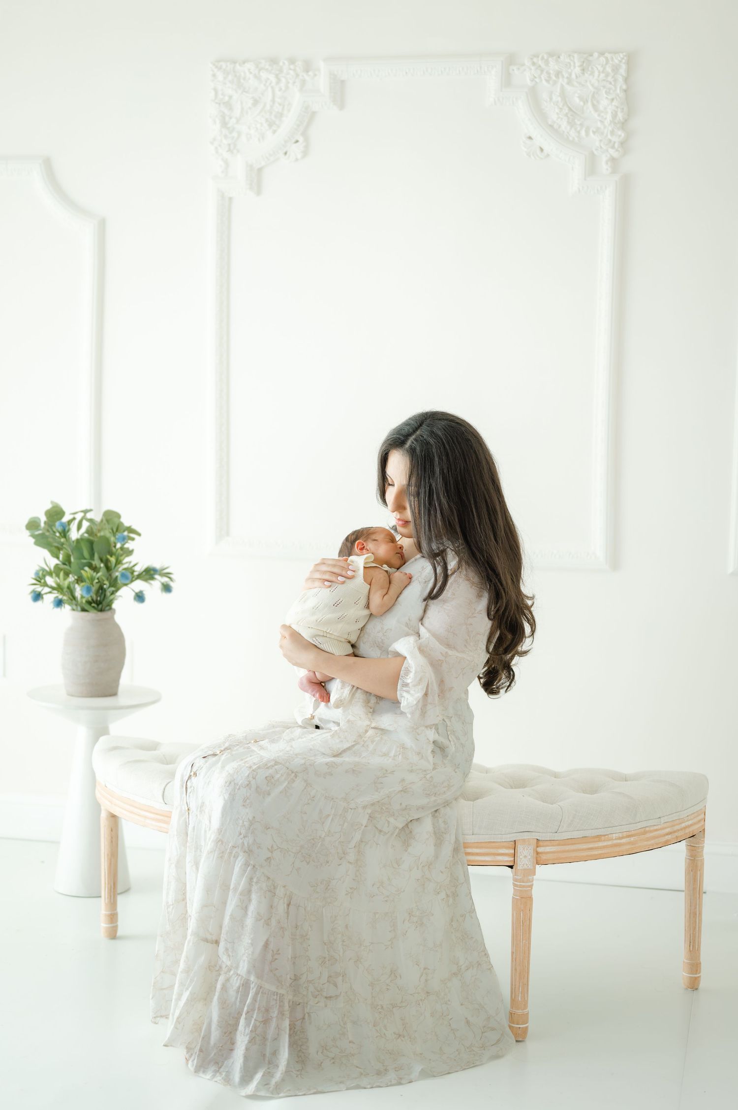 A mother in a lace gown sits on a bench cradling her newborn baby in a bright white elegant room.