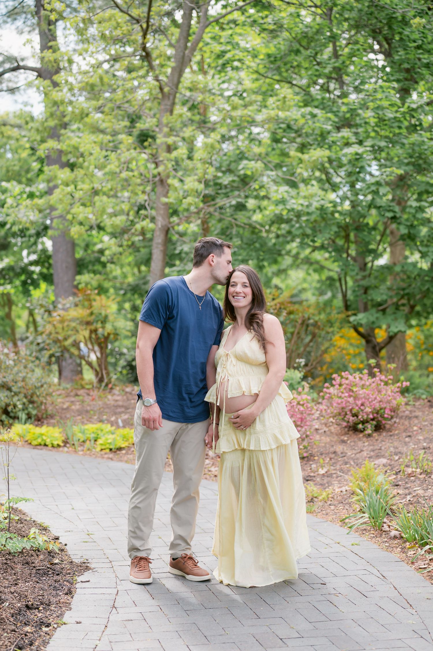 A man kisses a pregnant woman in a yellow dress on a garden path surrounded by lush greenery.