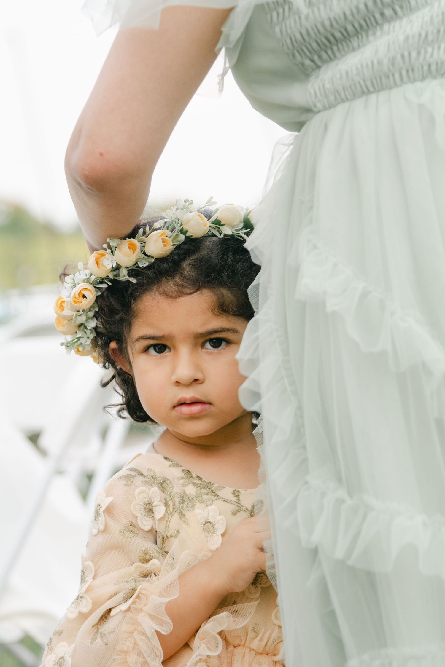 A small child wearing a floral crown and delicate cream-colored dress poses gracefully in a dreamy portrait setting.