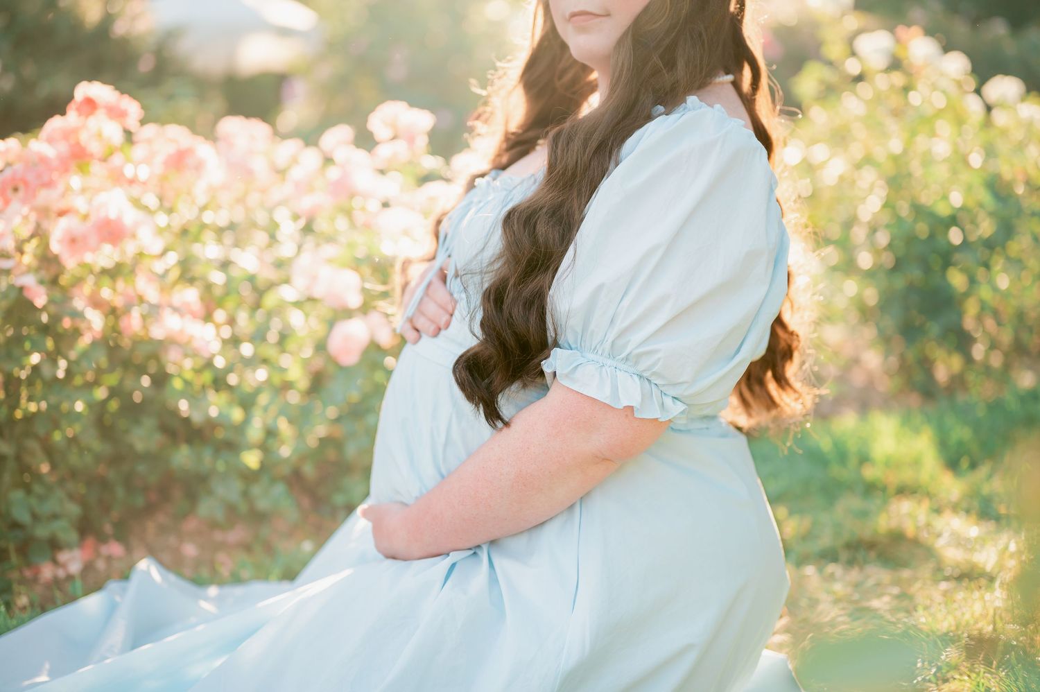 Pregnant woman in light blue dress cradling baby bump among blooming pink roses in soft golden sunlight.