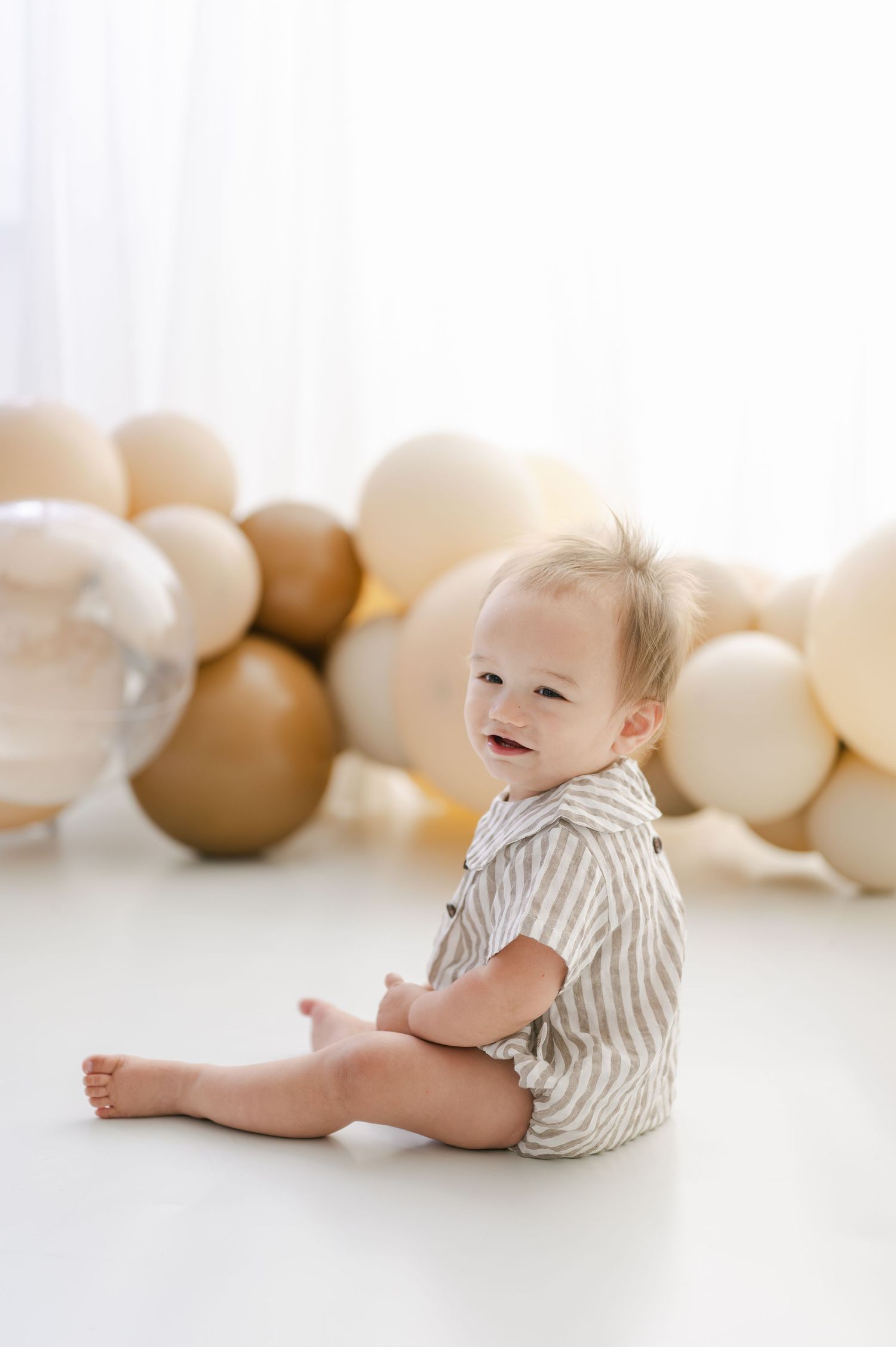 Smiling baby boy sitting on white floor surrounded by neutral beige and gold balloons in a bright studio setting.