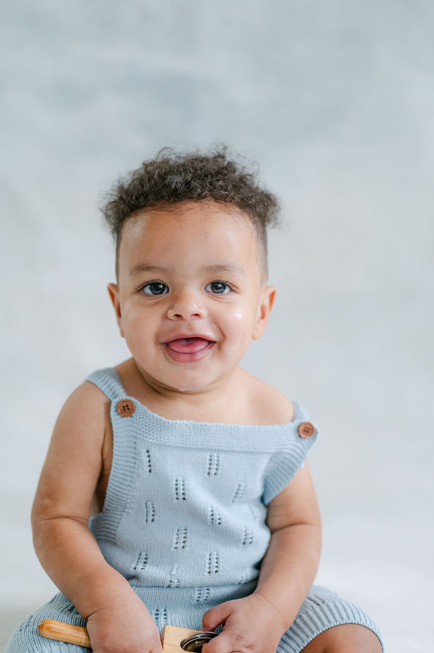 Smiling baby in a light blue knit romper with button straps, sitting against a soft gray background.