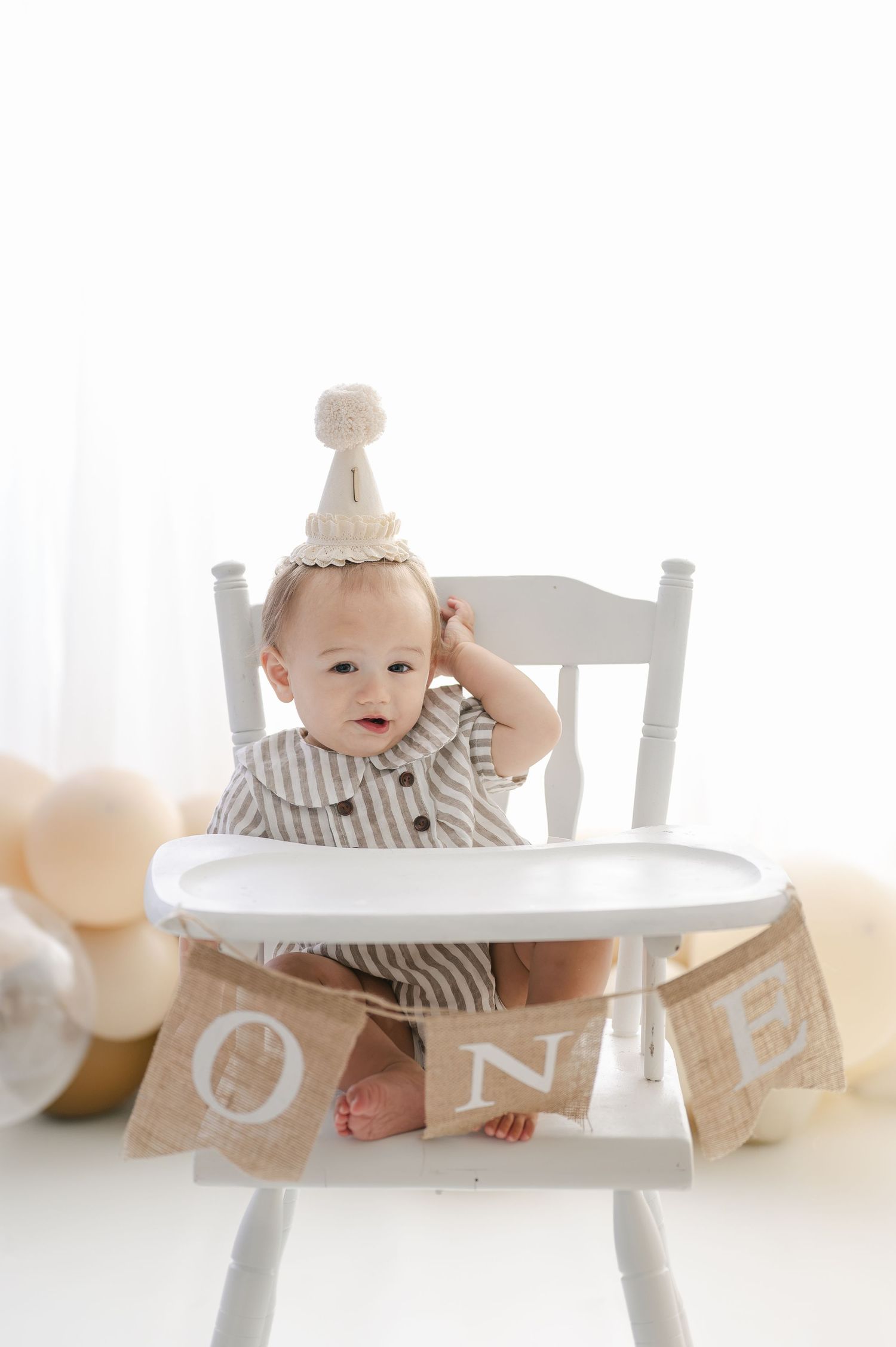 Baby in striped outfit and party hat sits in white high chair with ONE banner for first birthday cake smash photo.