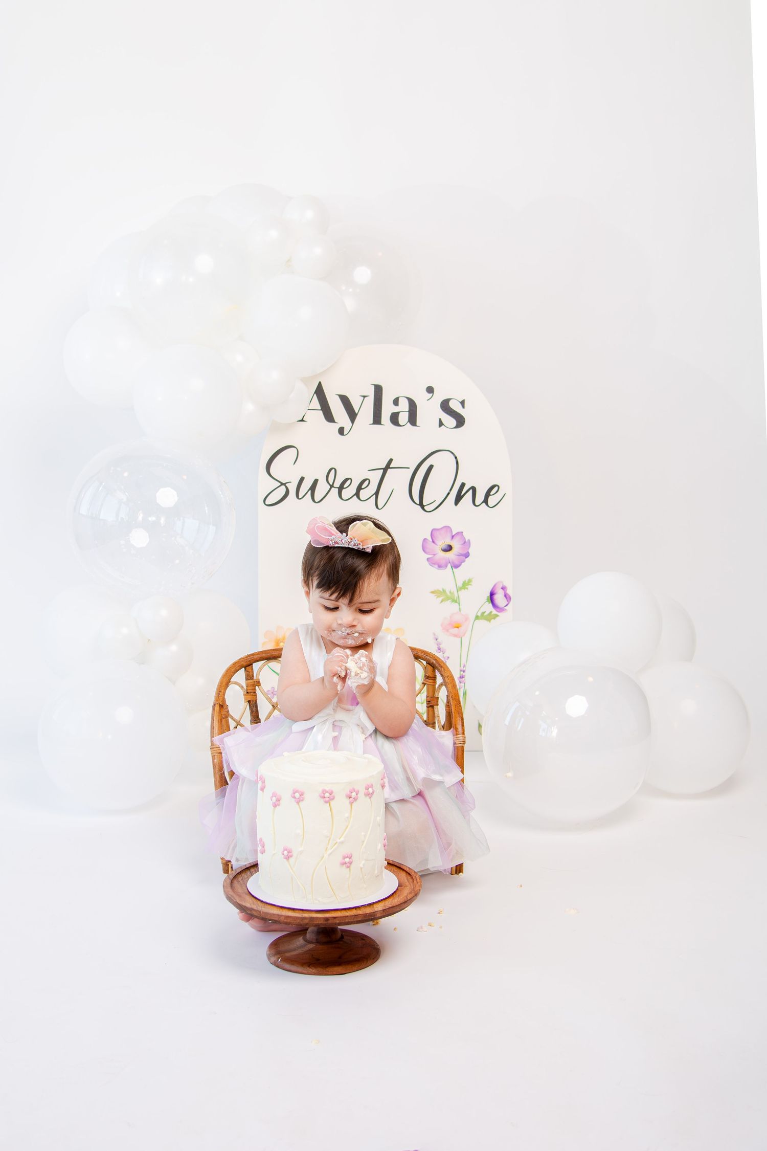Sweet pink and white birthday cake setup with white balloons on wooden surface for first birthday celebration.