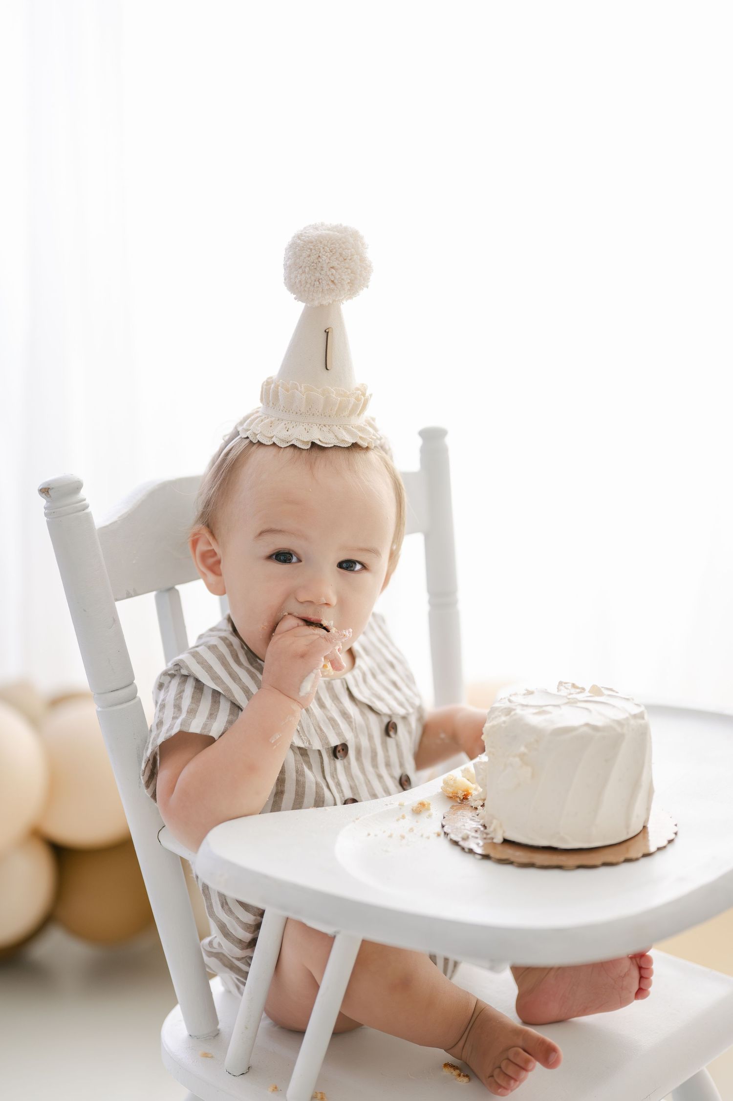 Baby in a white high chair wearing a birthday hat eating a smash cake during a first birthday photo session.