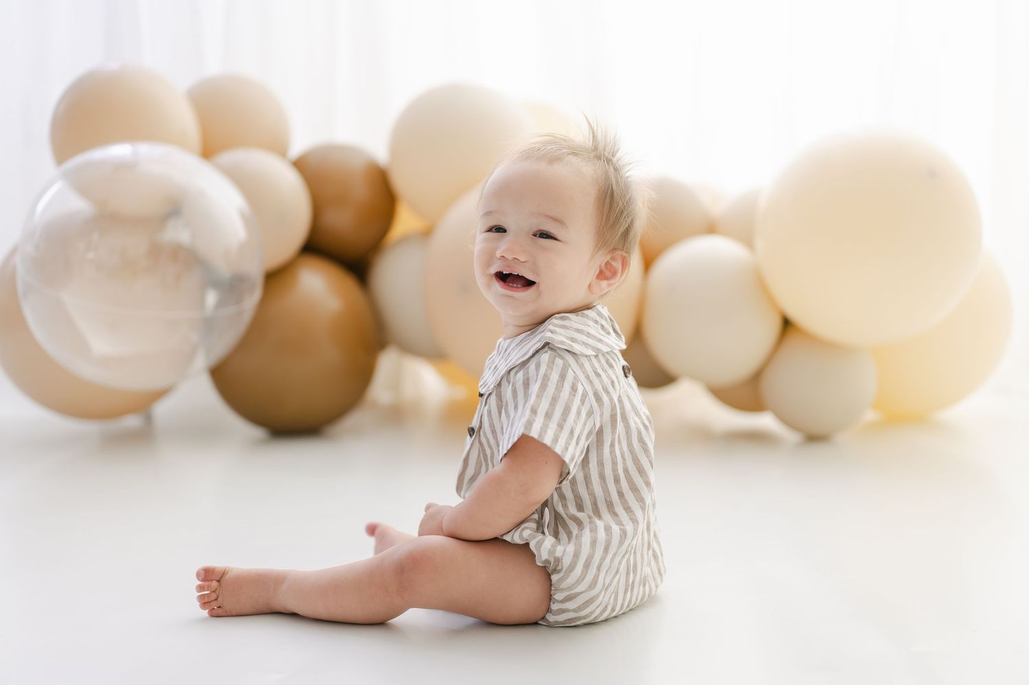 Smiling baby in striped romper sits on white floor surrounded by gold, cream, and clear balloons.
