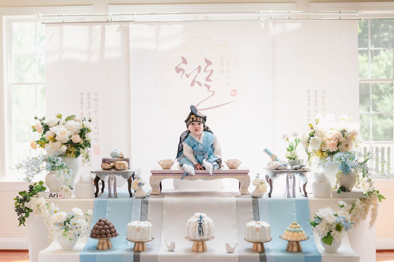 Child in traditional Asian attire sits at a decorated table surrounded by floral arrangements and elegant desserts at a celebration.