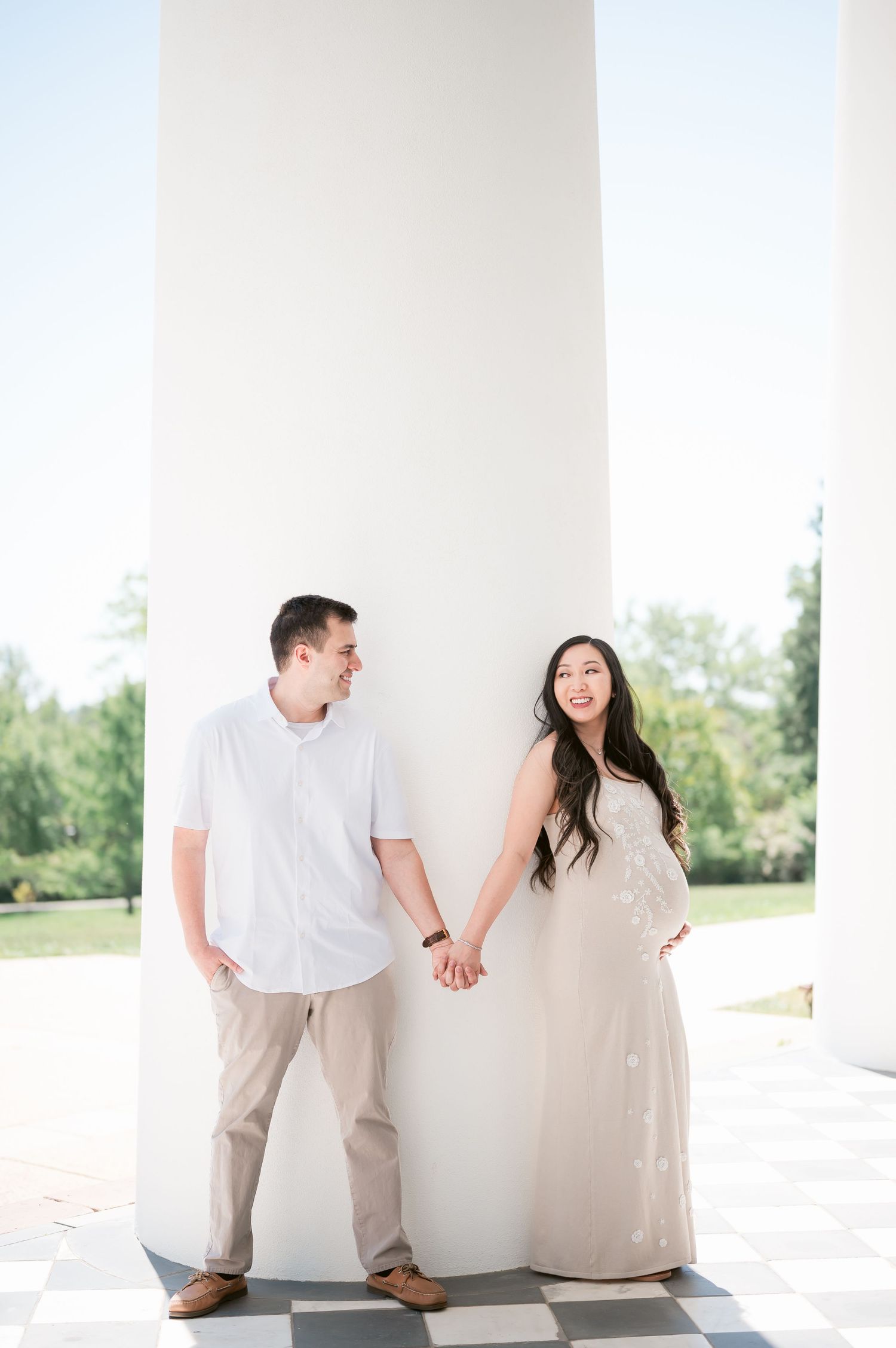 Couple holding hands in white outfits leaning against large white columns outdoors during a bright sunny day.