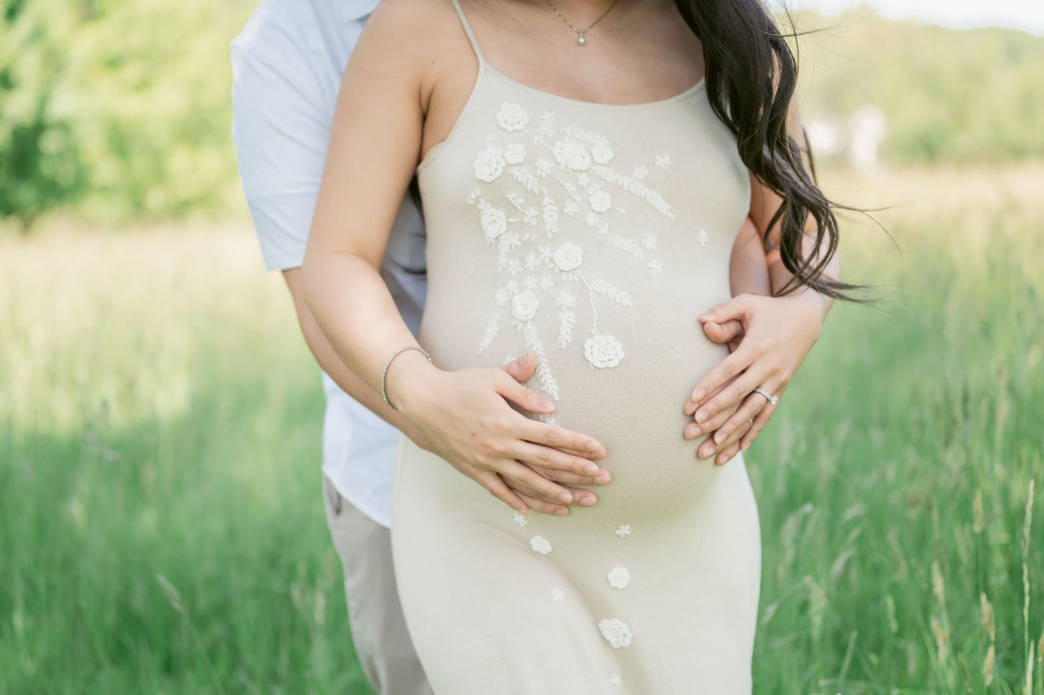 Couple in a green field, man embracing pregnant woman in a white floral dress during a maternity photo shoot.