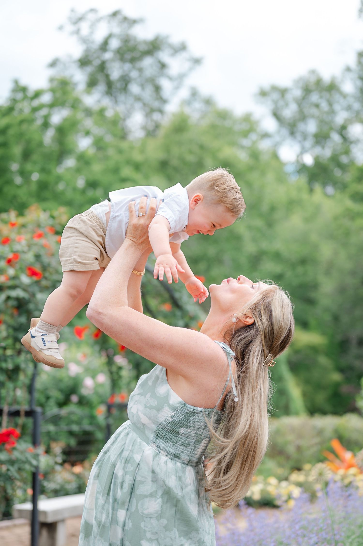 A joyful mother lifts her laughing baby boy overhead in a lush garden filled with colorful blooming flowers.