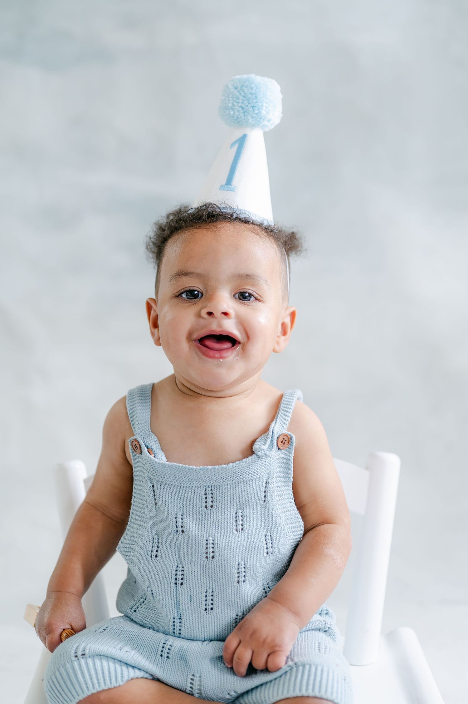 Smiling baby in blue knit romper wearing a number one birthday hat sits in a white chair for first birthday photos.