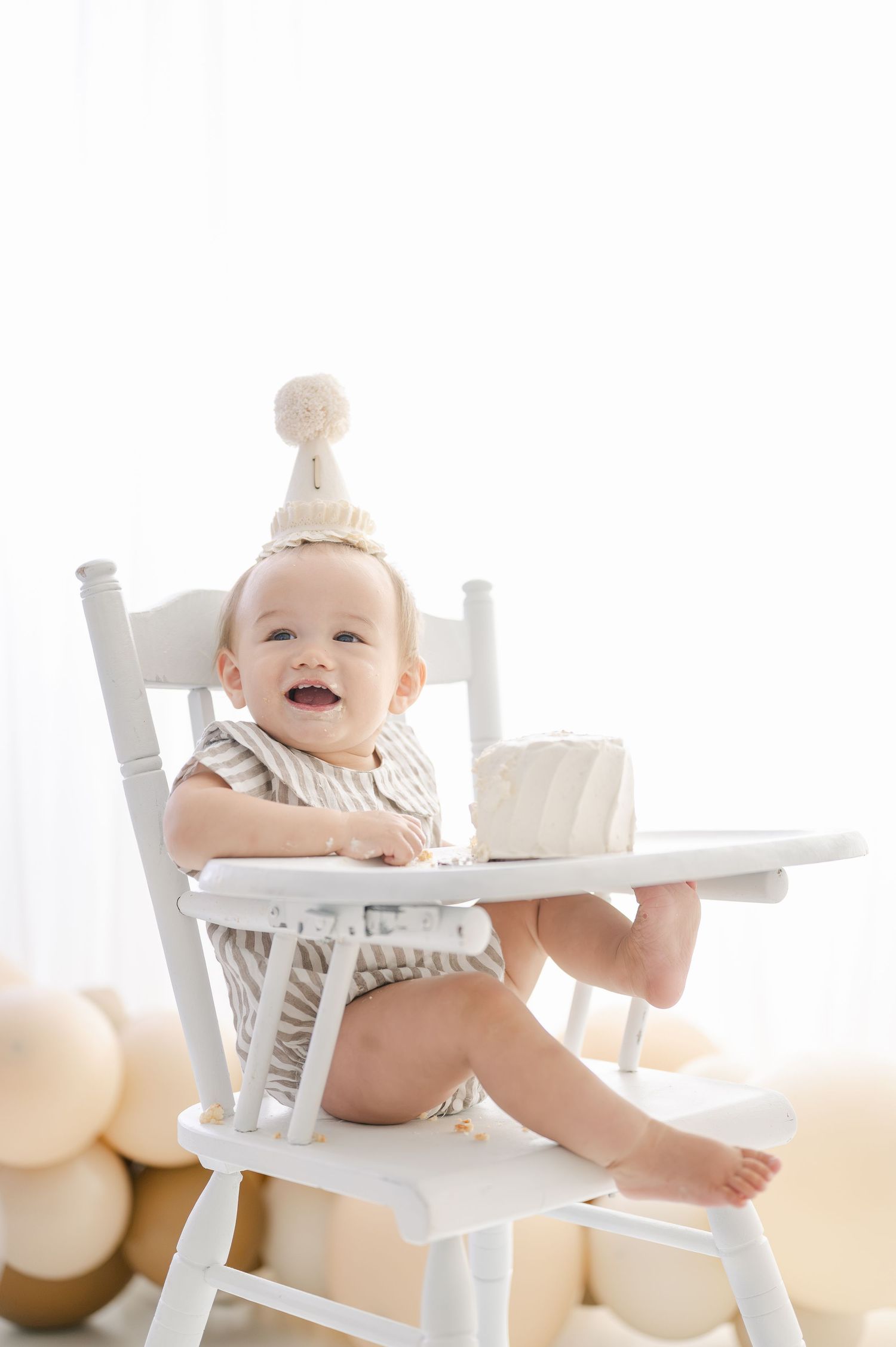 Smiling baby in white high chair wearing birthday hat with cake, celebrating first birthday in neutral beige themed photo.