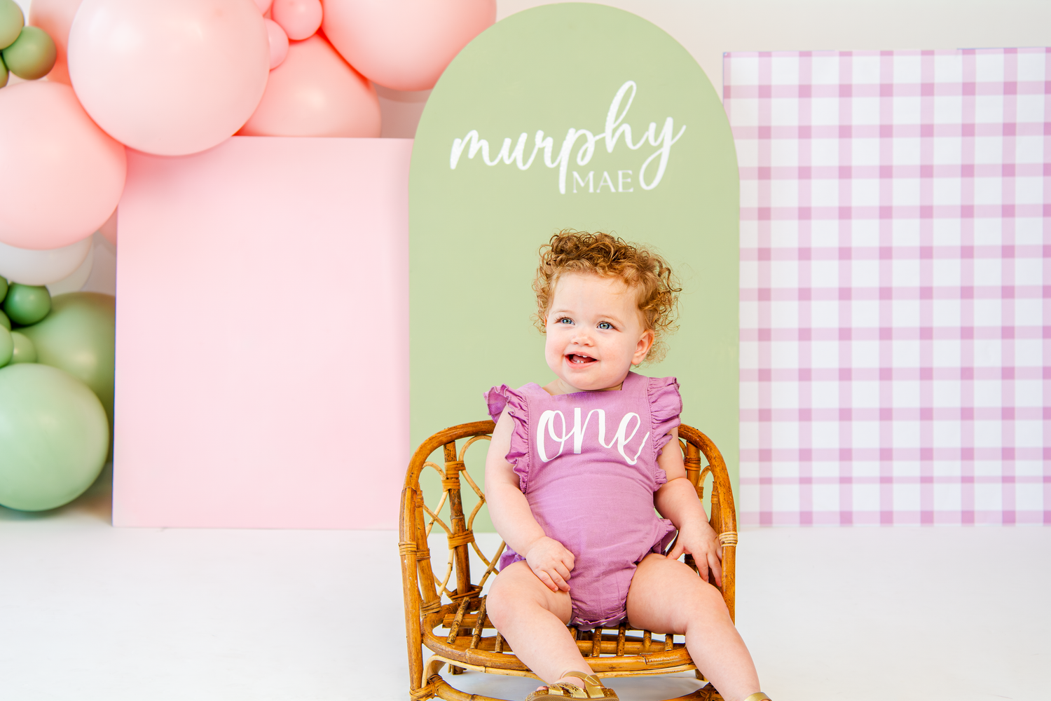 First birthday photo setup featuring pink and green balloons with gingham pattern background.