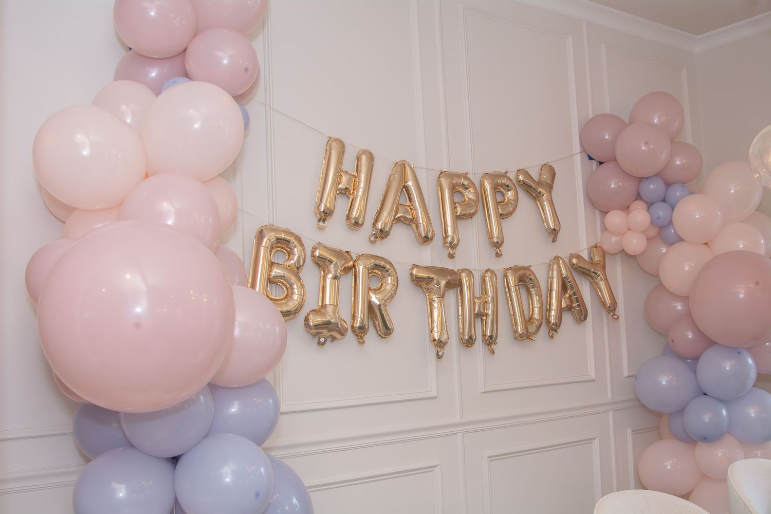 Pink and purple balloon garland with gold 'HAPPY BIRTHDAY' letter balloons decorates a white wall for a celebration.