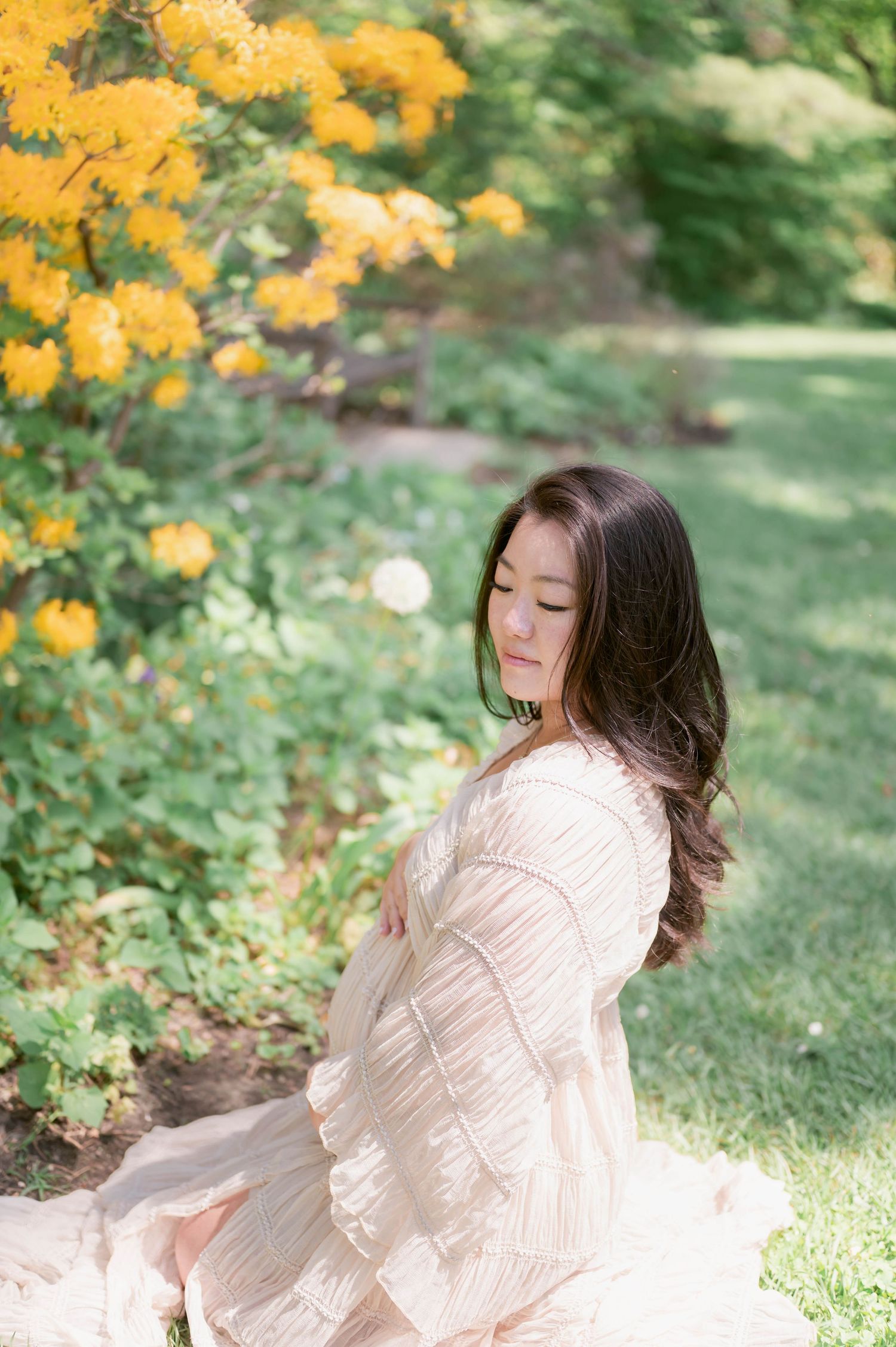 Pregnant woman in blush dress sitting outdoors near yellow flowers, cradling her baby bump in a lush green garden.