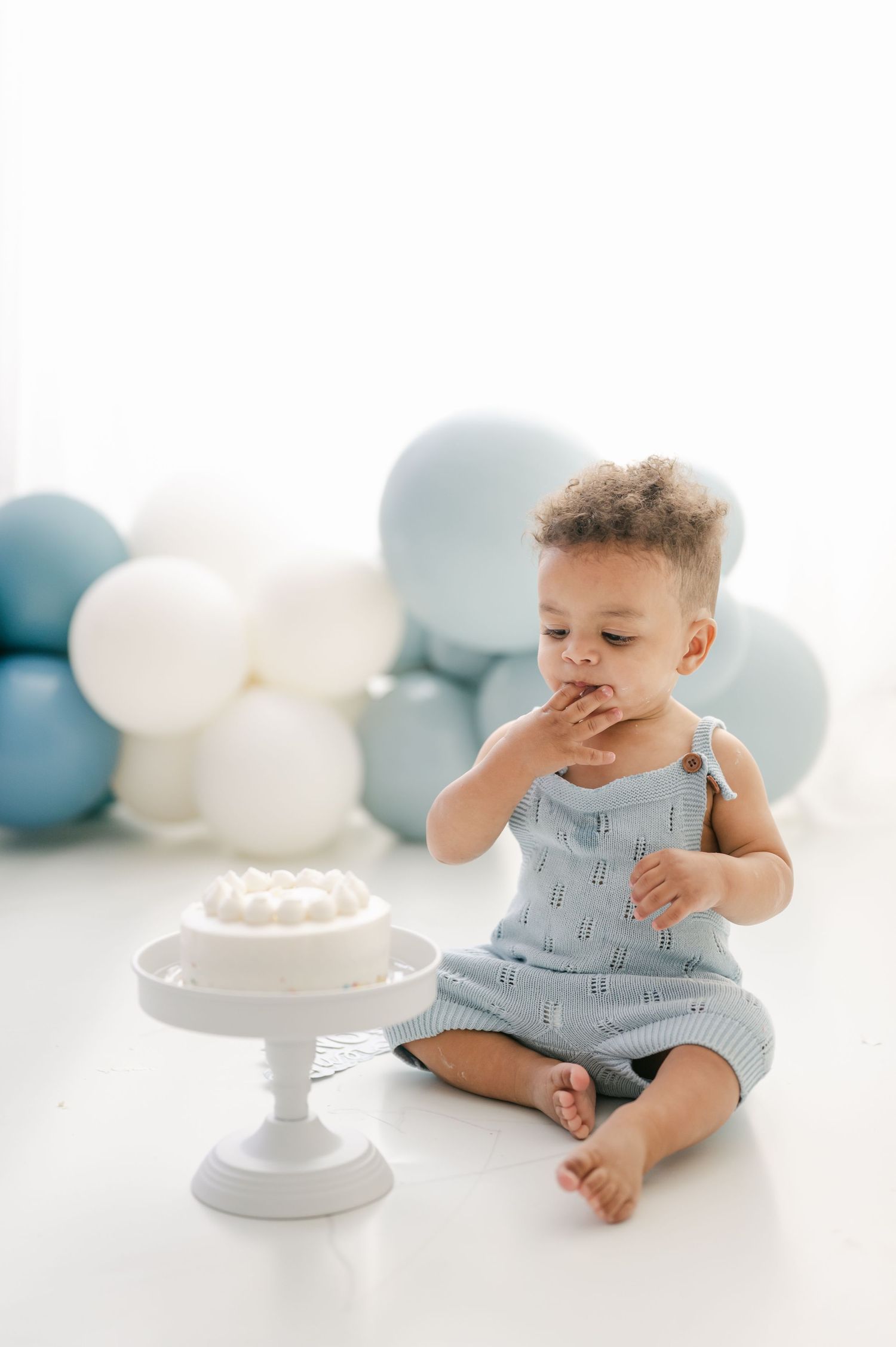 A baby boy in a blue romper sits on a white floor, eating cake at his first birthday smash cake photoshoot.