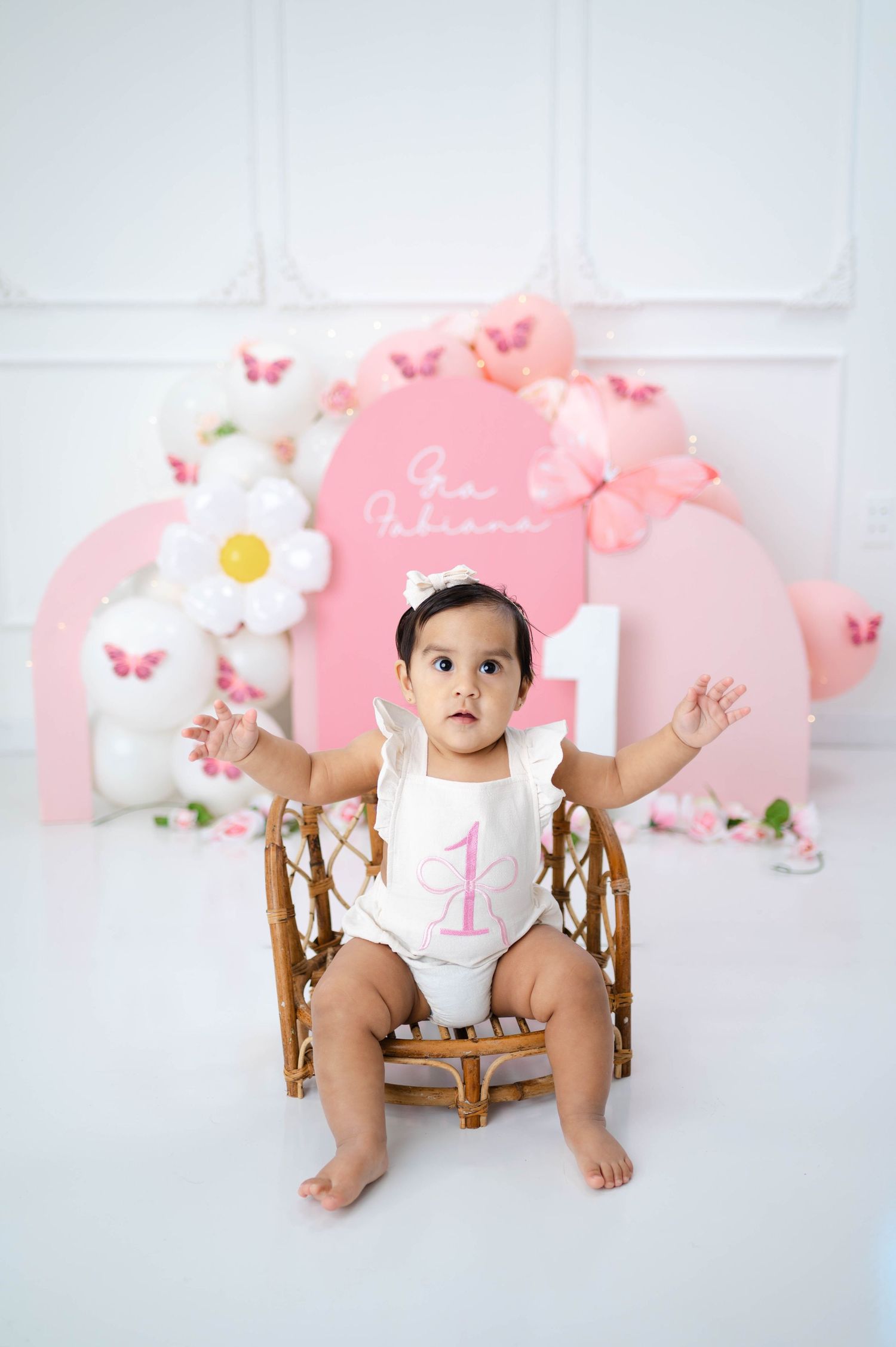 Pink and white themed first birthday photo setup with butterflies and floral decorations against white backdrop.