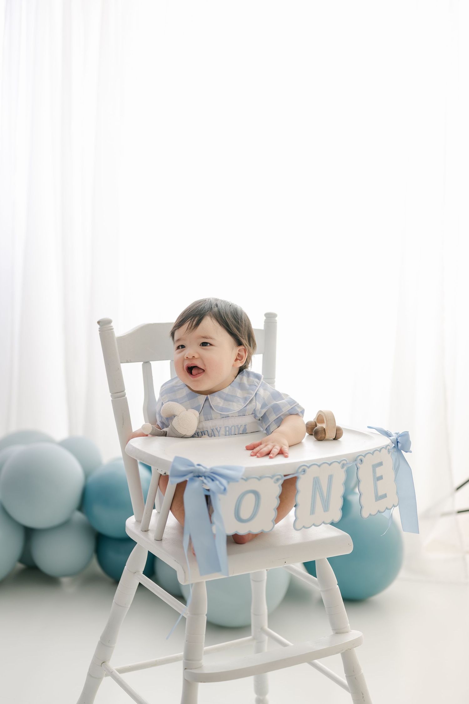 Baby in white high chair with ONE banner and blue balloons celebrates first birthday.