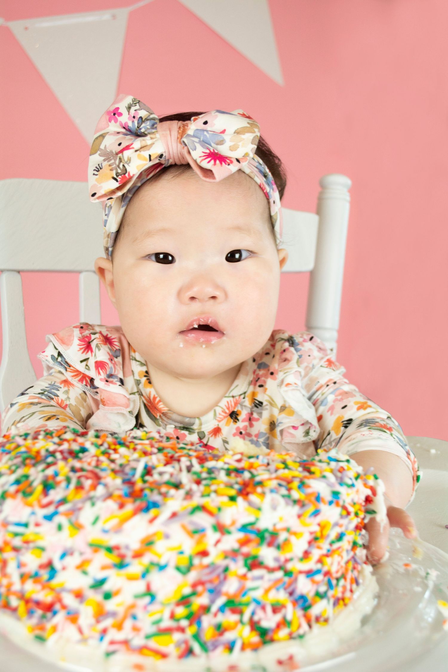 Baby wearing floral headband explores colorful sprinkle-covered cake during pink-themed photoshoot.