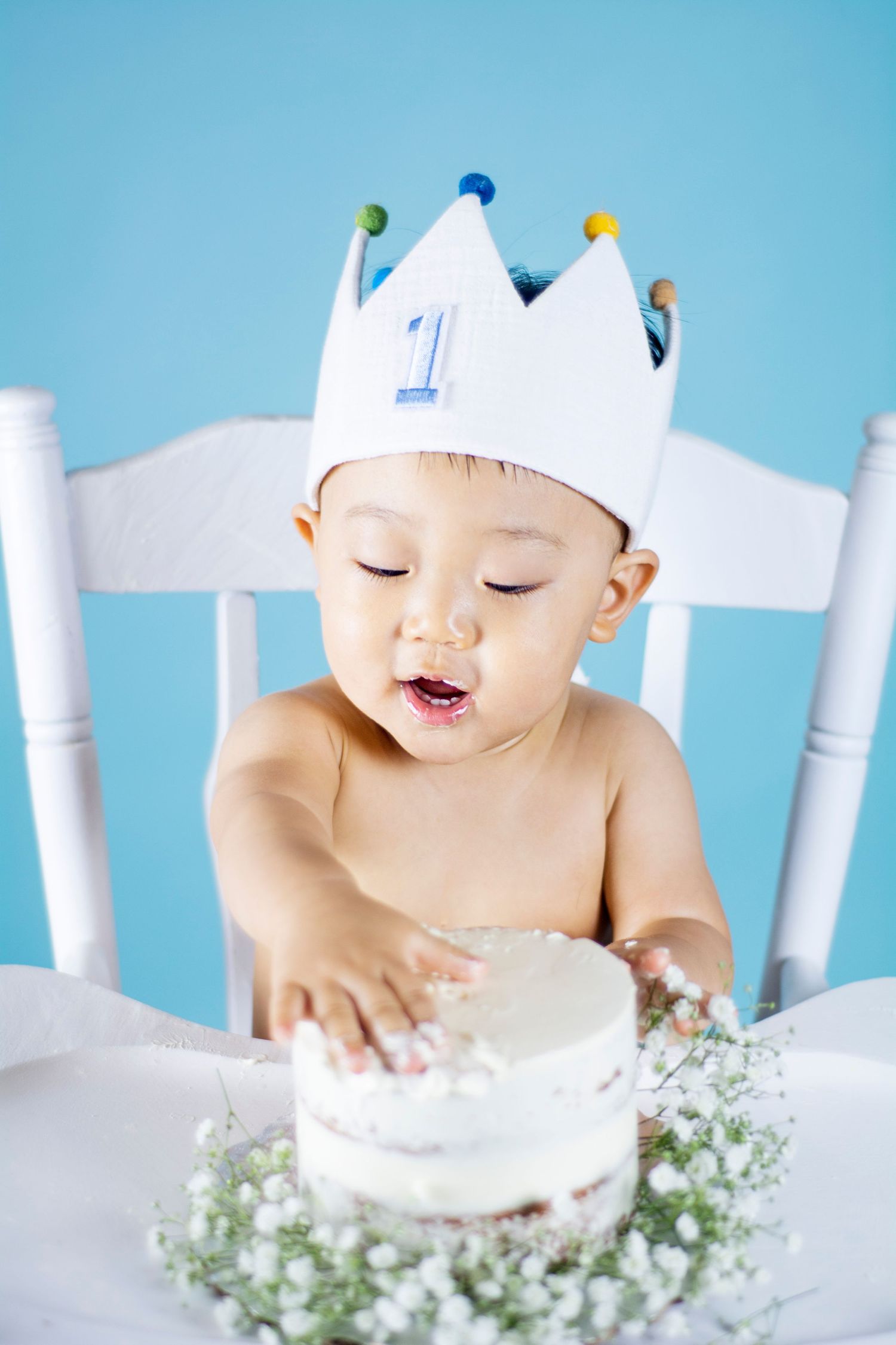 Baby wearing paper crown leans over white cake during first birthday photoshoot on blue backdrop.