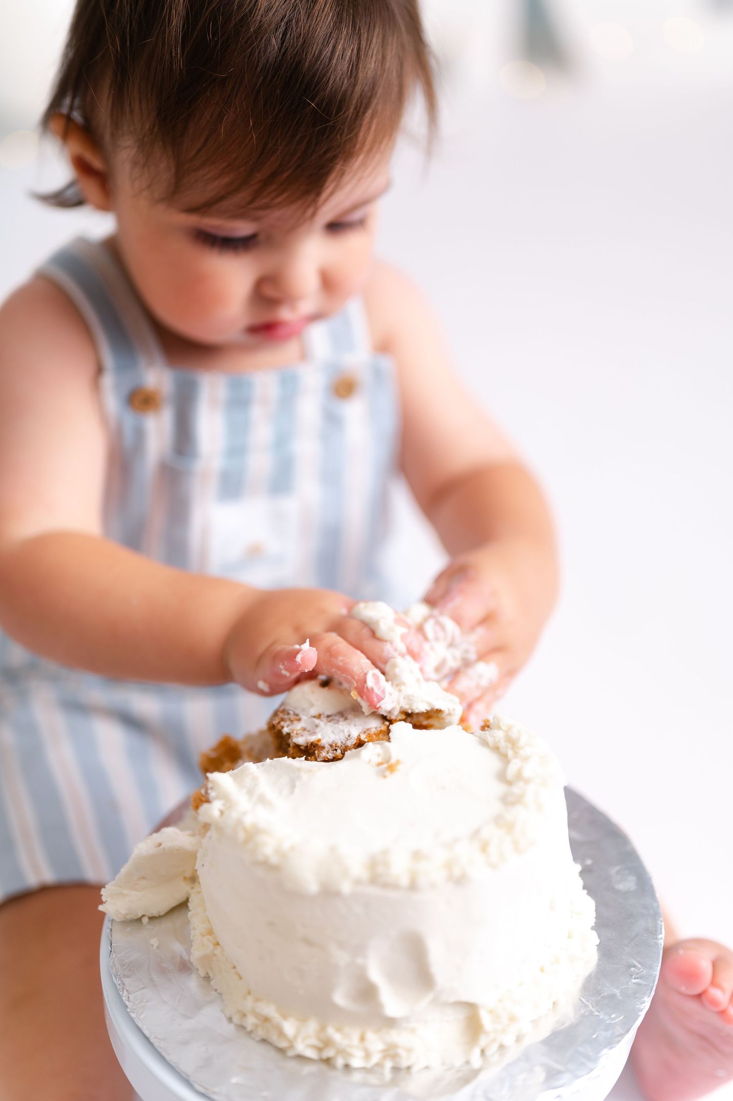 A baby in a blue striped romper smashes their first birthday cake with white frosting during a cake smash photo session.
