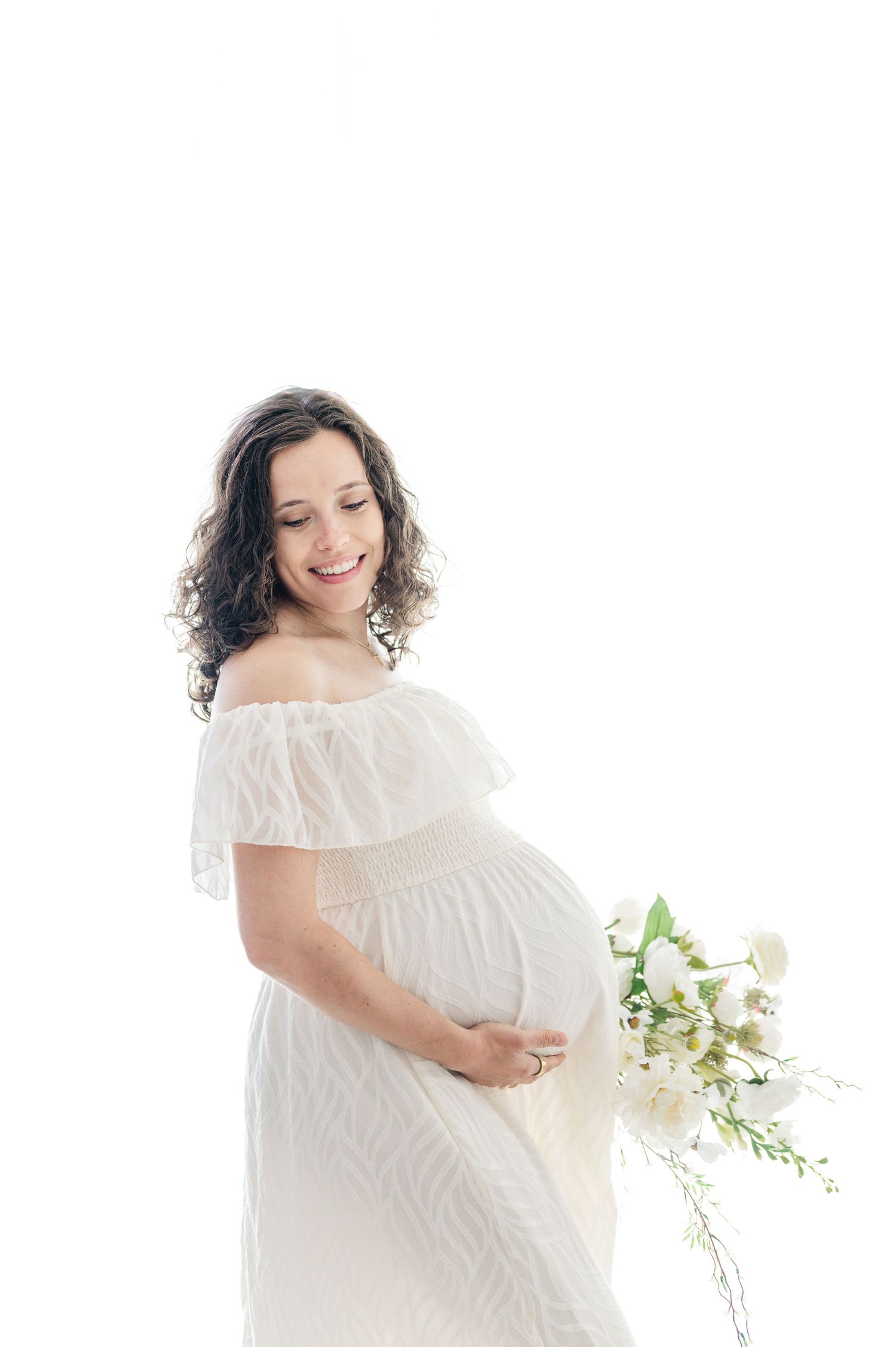 Smiling pregnant woman in white off-shoulder dress holding white flowers, gently cradling her baby bump against white background.