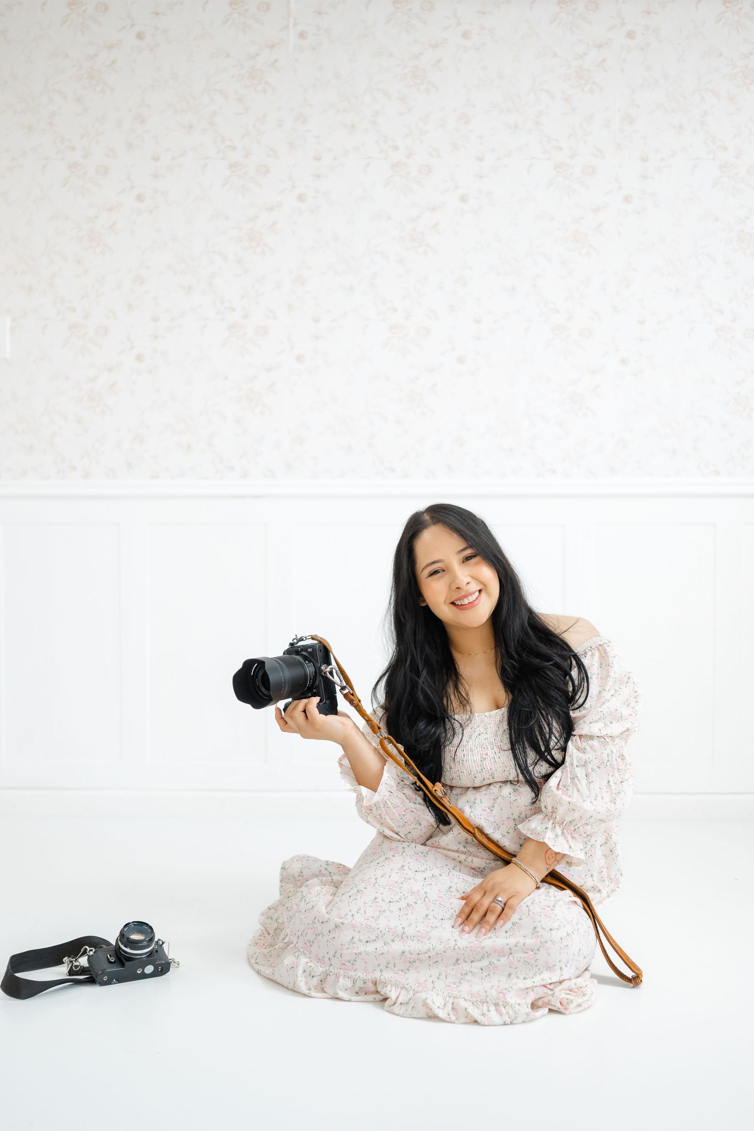 Smiling Asian woman in floral dress sitting on white floor holding DSLR camera with second camera beside her.