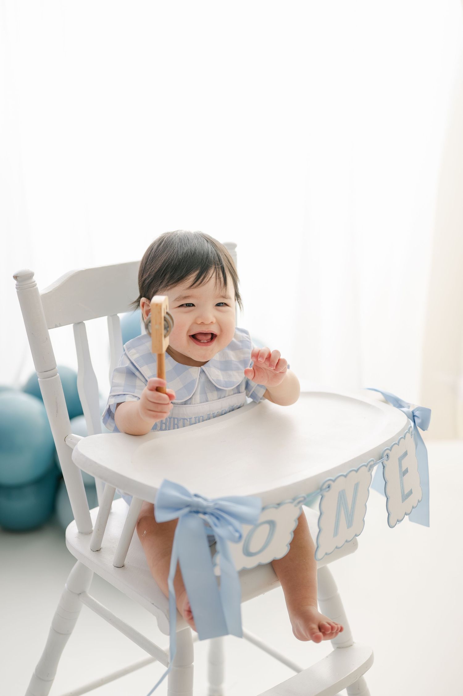 A happy baby sits in a white high chair holding a wooden spoon, celebrating their first birthday with a blue bow banner.