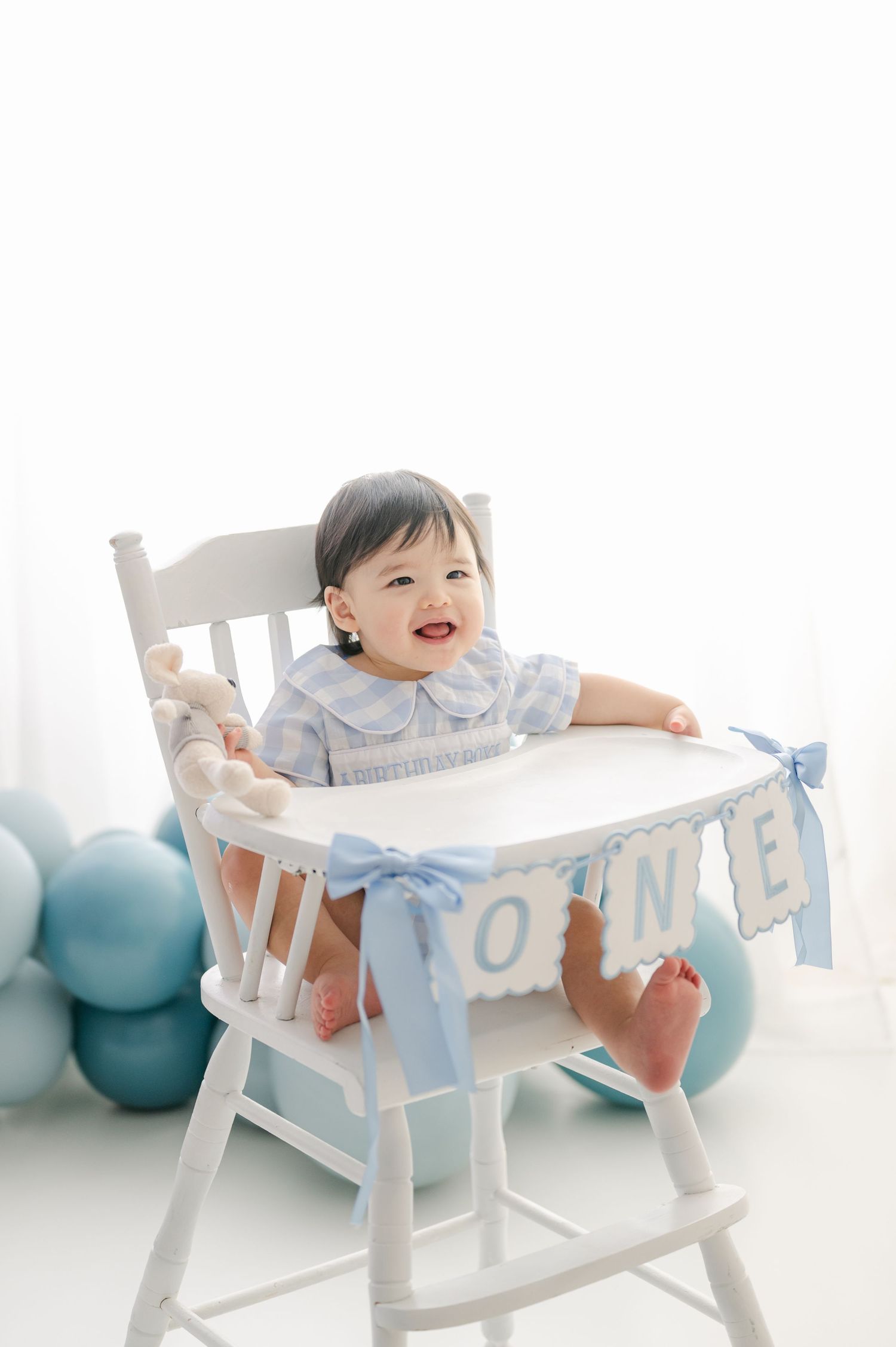 Smiling baby in blue outfit sits in white high chair decorated with ONE banner and blue balloons for first birthday.