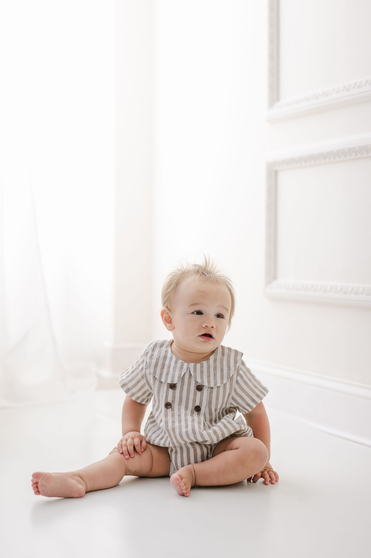 Cute baby sitting on white floor wearing striped romper in bright white studio setting.
