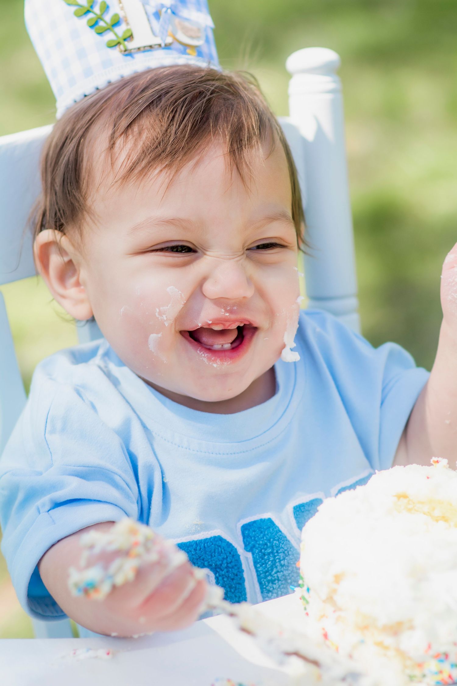 Baby enjoying a cake smash photoshoot seated in blue high chair outdoors.