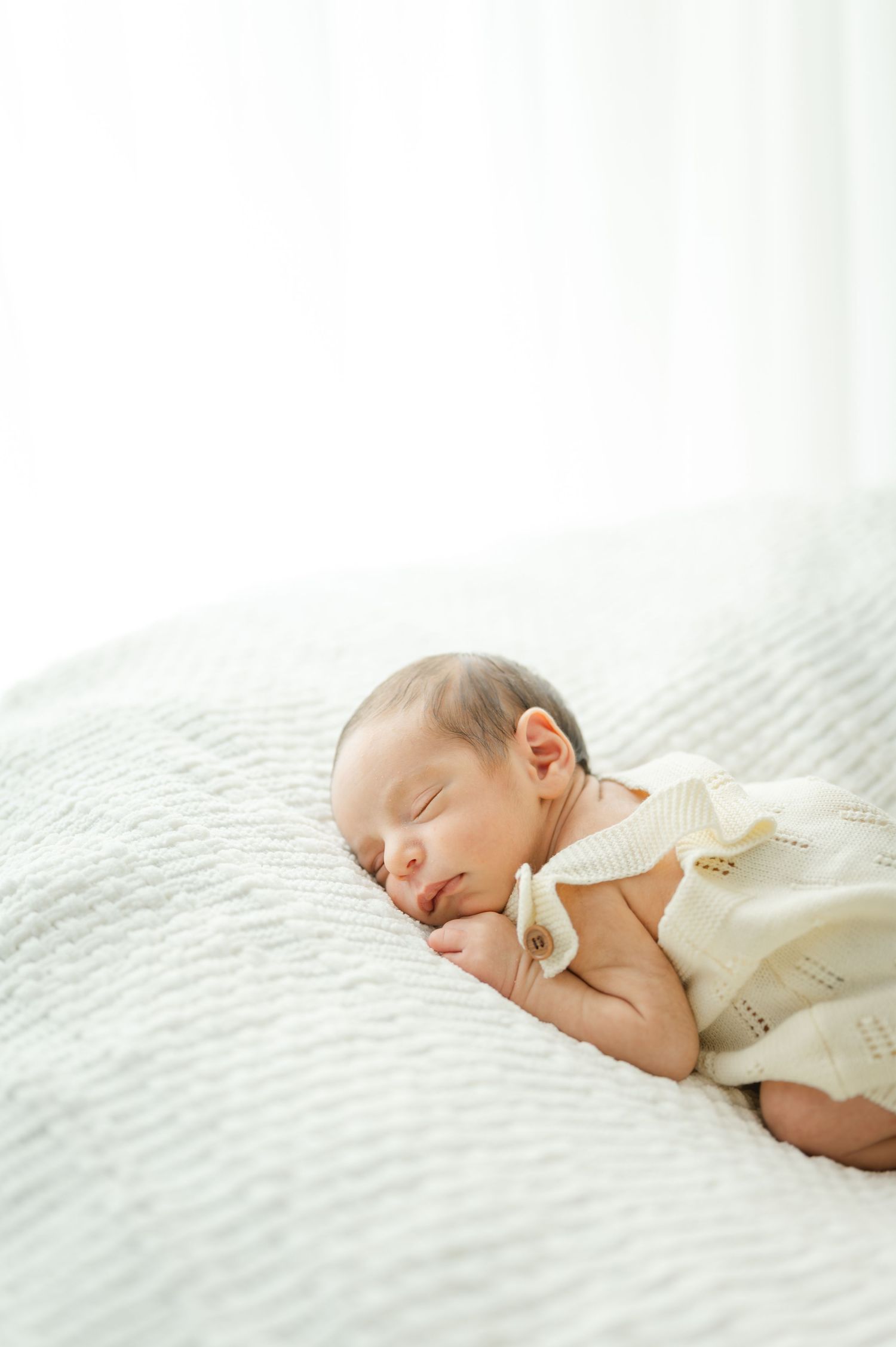 Peaceful newborn baby sleeping on white quilted blanket wearing cream knit romper in soft natural light.