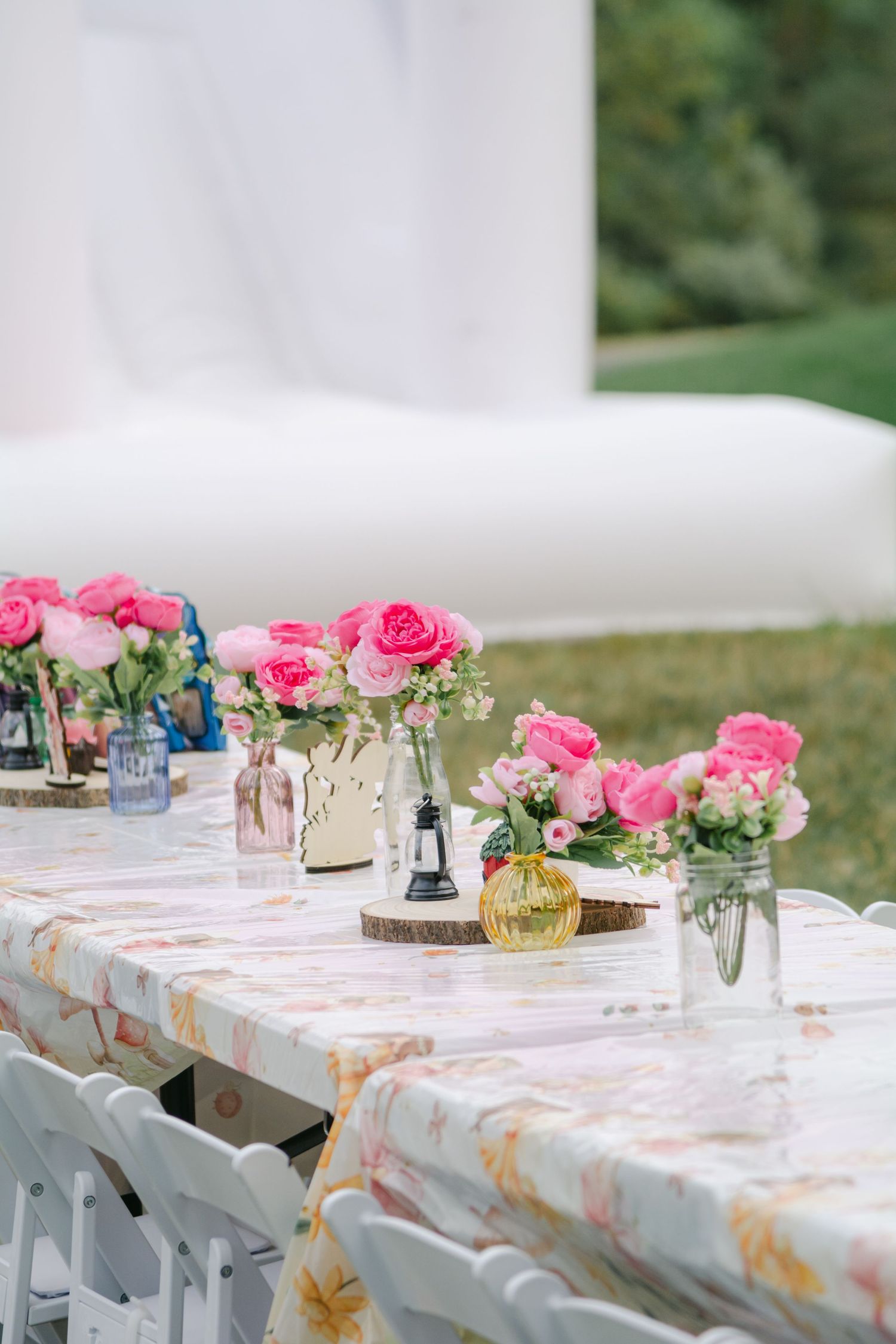 Long white tables adorned with pink roses and rustic wood slices create an elegant outdoor dining setup.