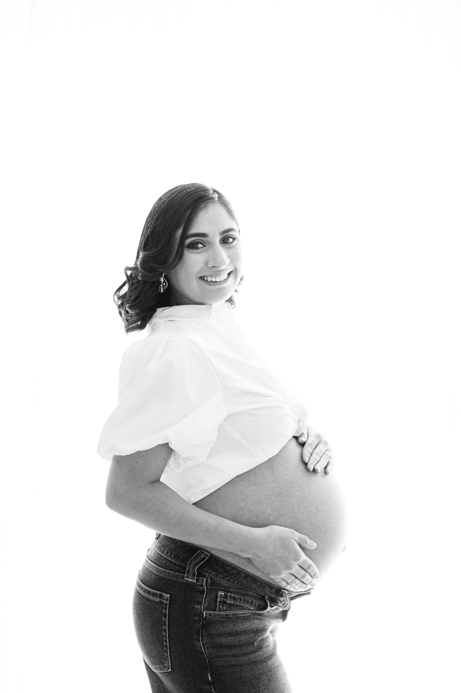 Black and white maternity photo of a smiling pregnant woman in white top and jeans cradling her baby bump.