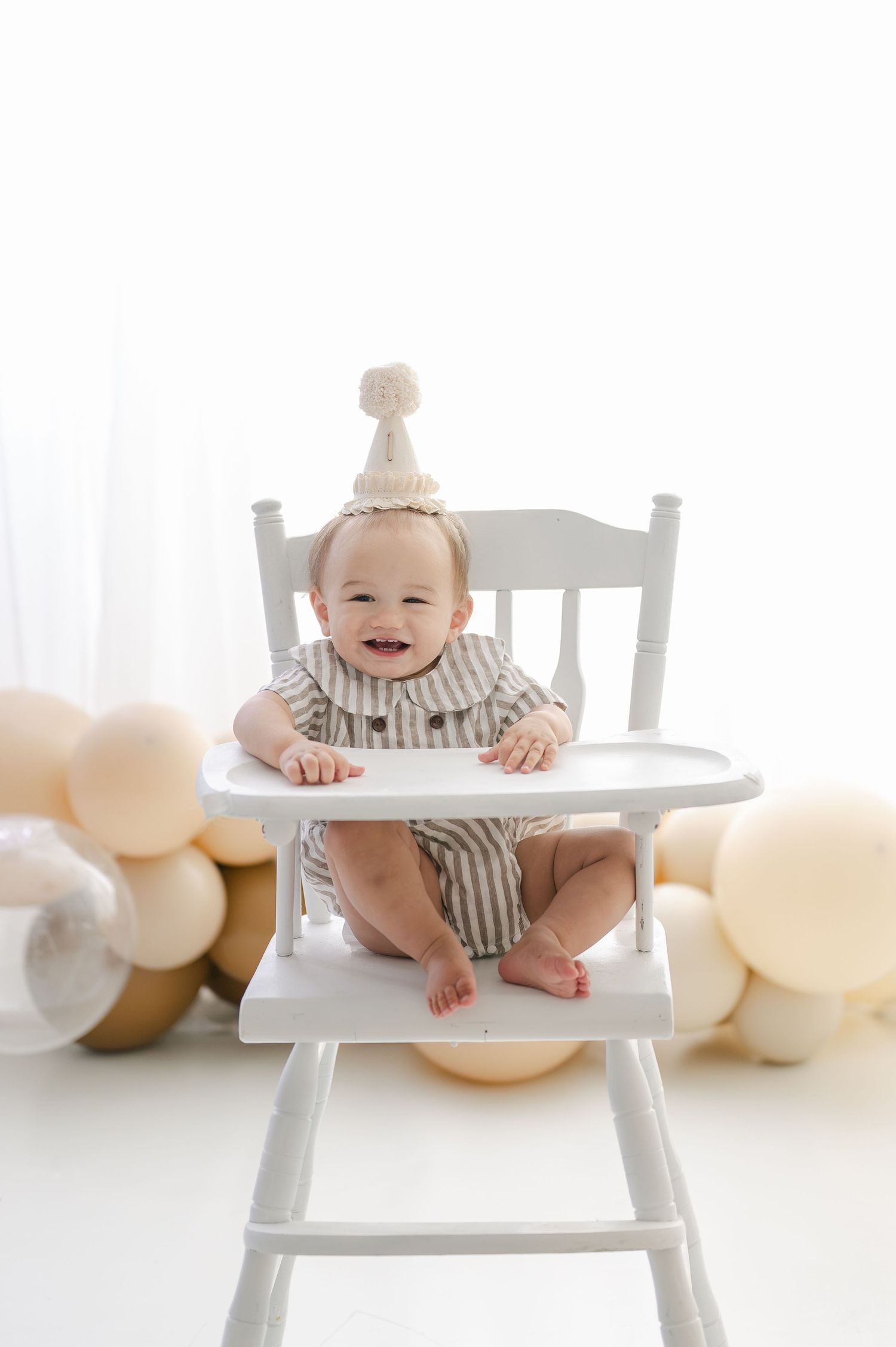 Smiling baby in striped romper and party hat sits in white high chair surrounded by gold and cream balloons.