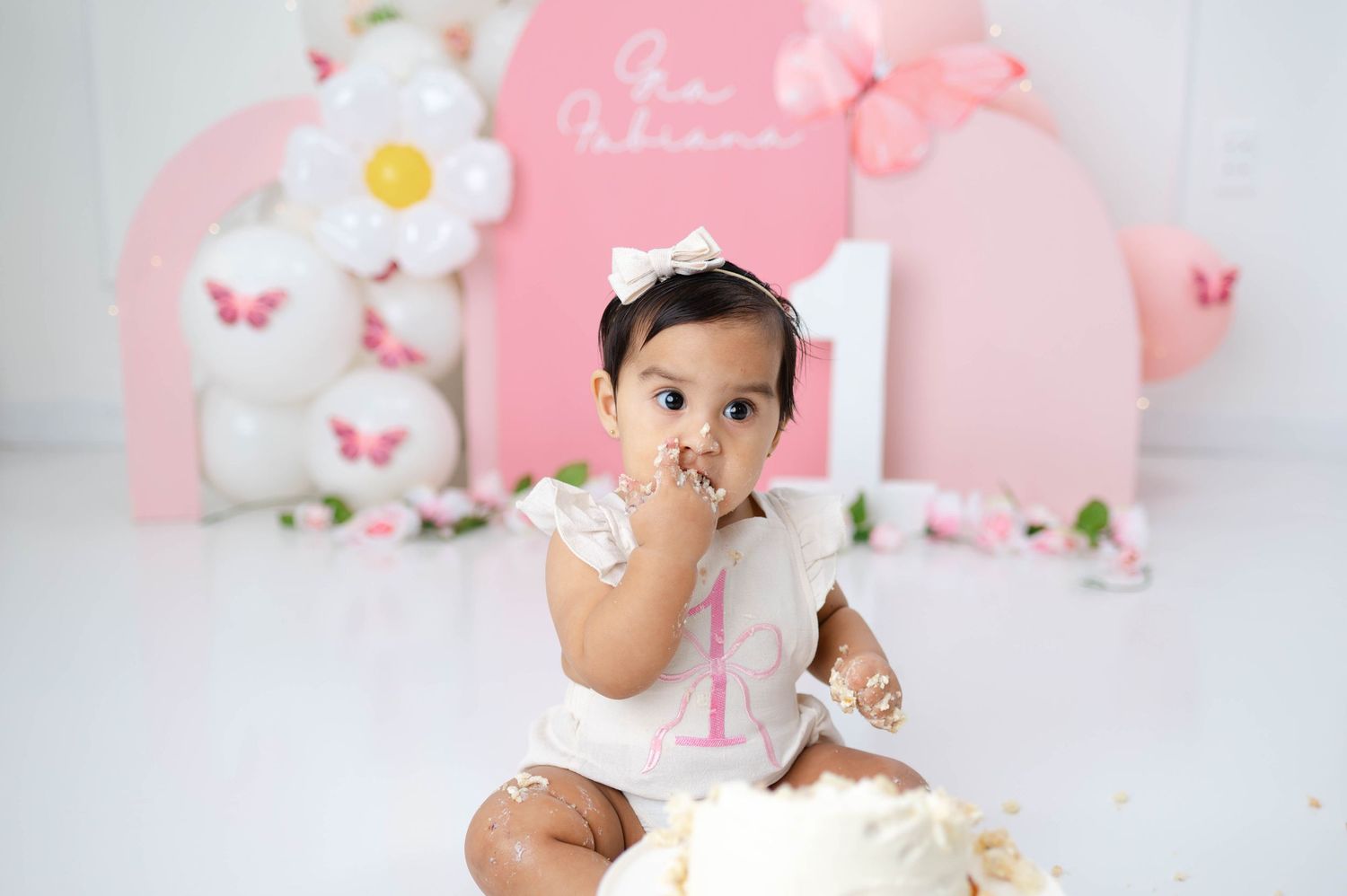 Pink and white themed cake smash photo session setup with butterflies and floral decorations for a first birthday celebration.