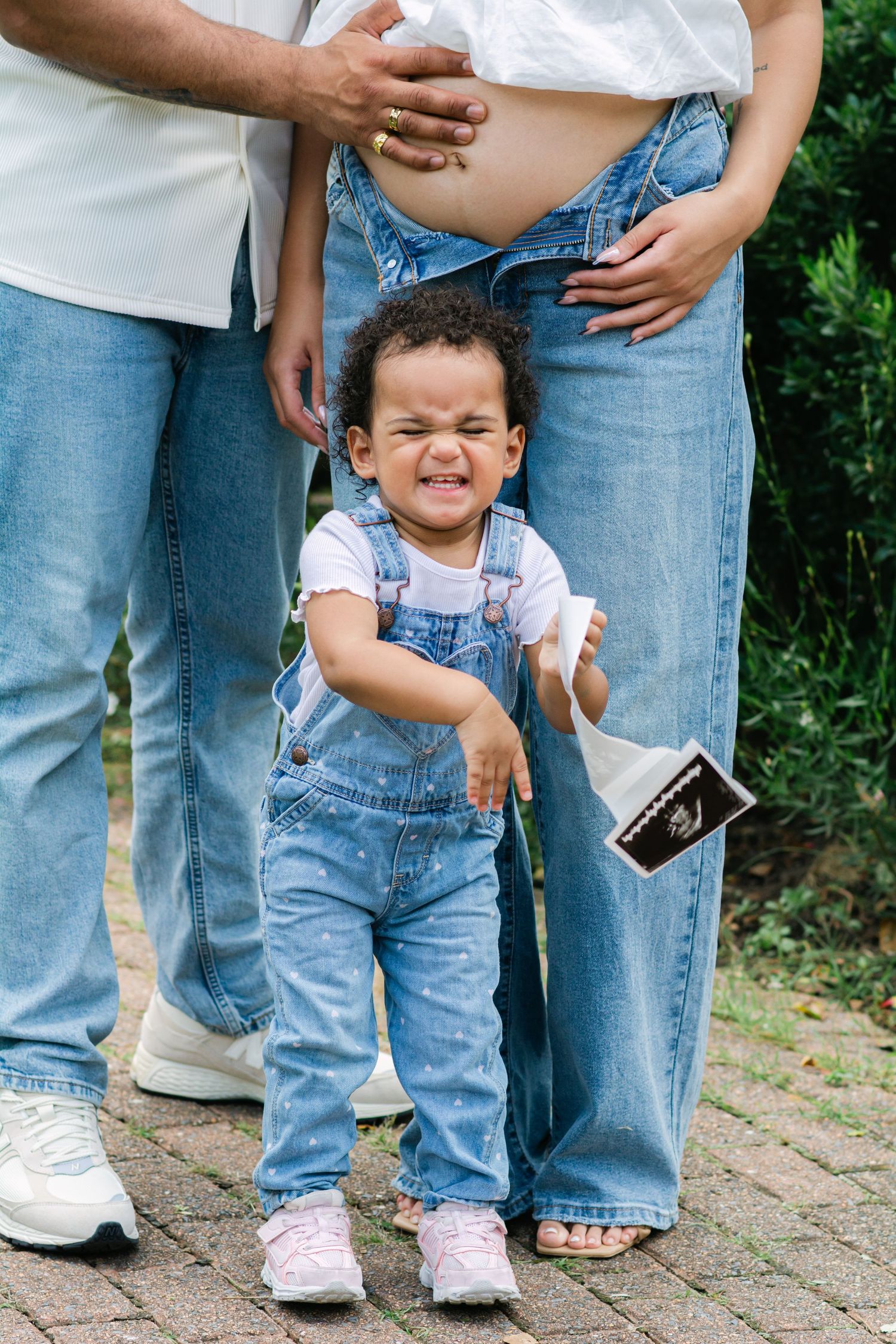A small child in denim overalls plays near adults holding ultrasound photos in an outdoor setting.