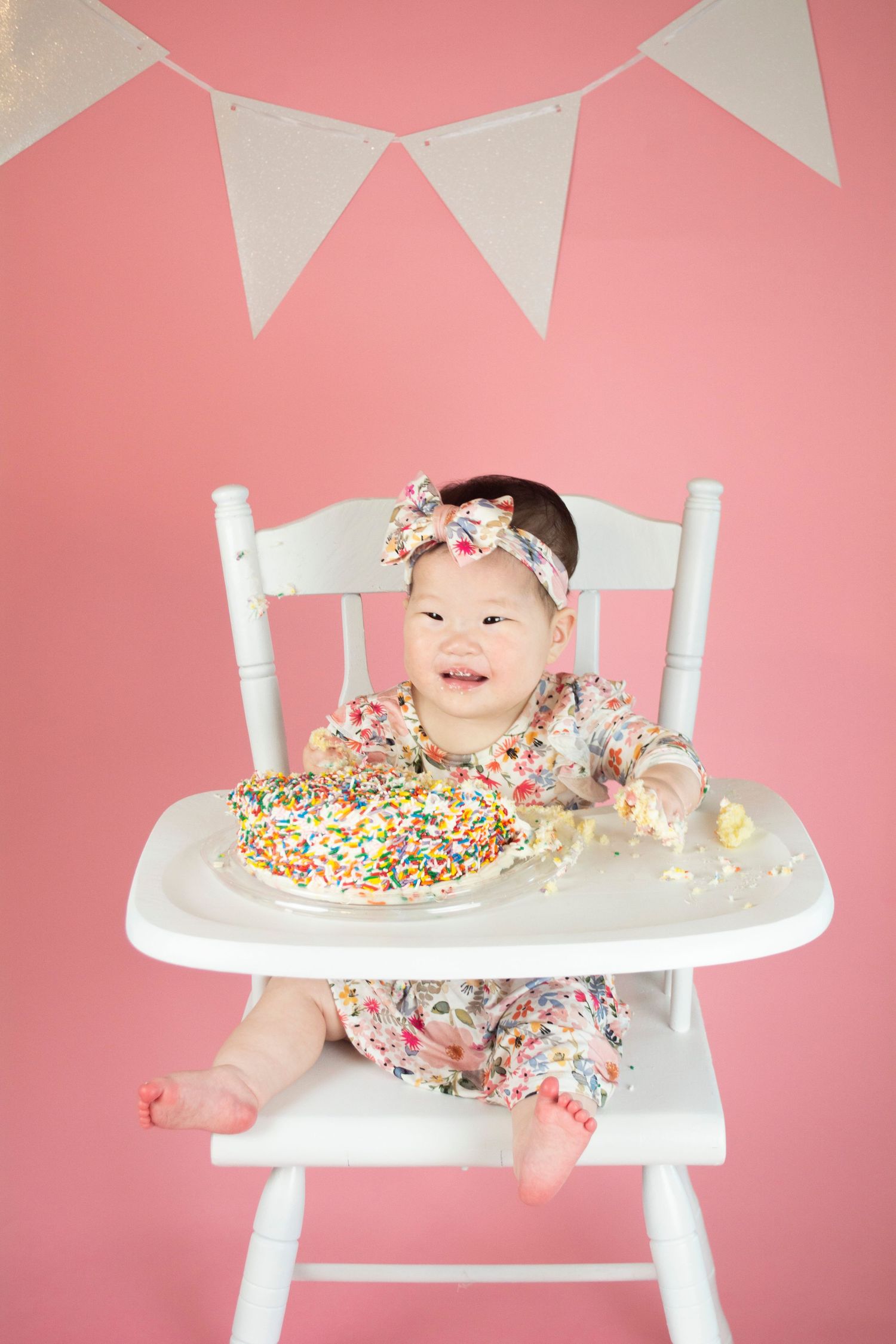 Baby in floral outfit sits in white high chair with cake during pink-themed first birthday shoot.