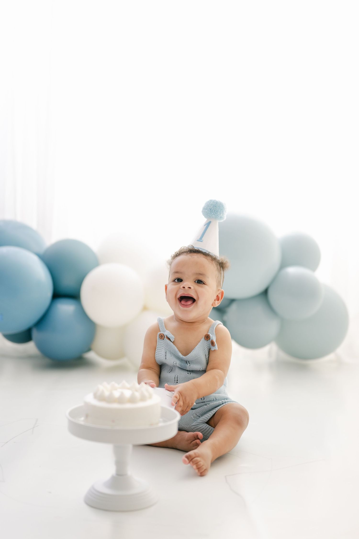 Happy baby in blue romper and party hat sits by smash cake, surrounded by blue and white balloons.