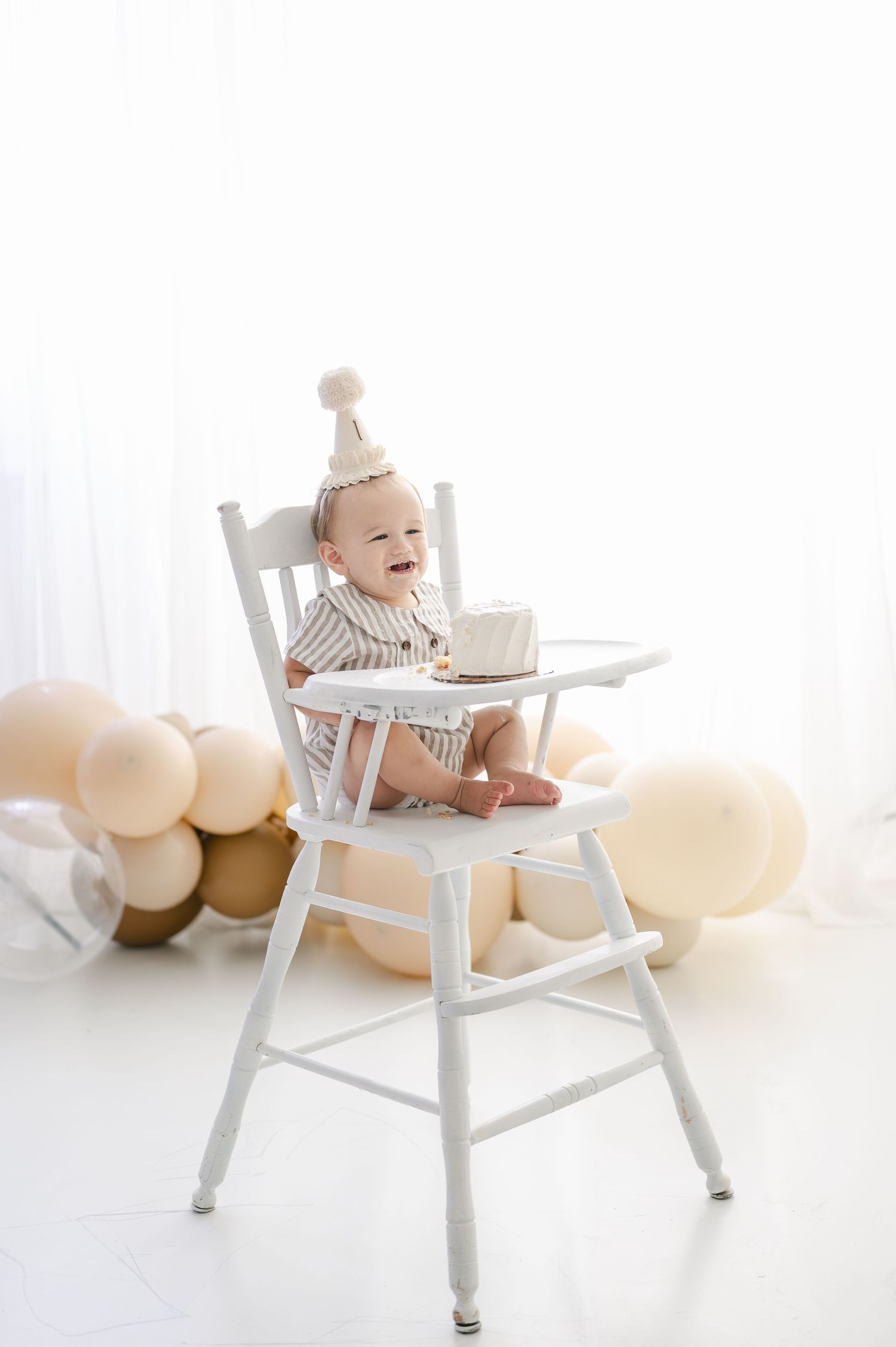 Happy baby in white high chair wearing party hat surrounded by beige balloons celebrating first birthday.