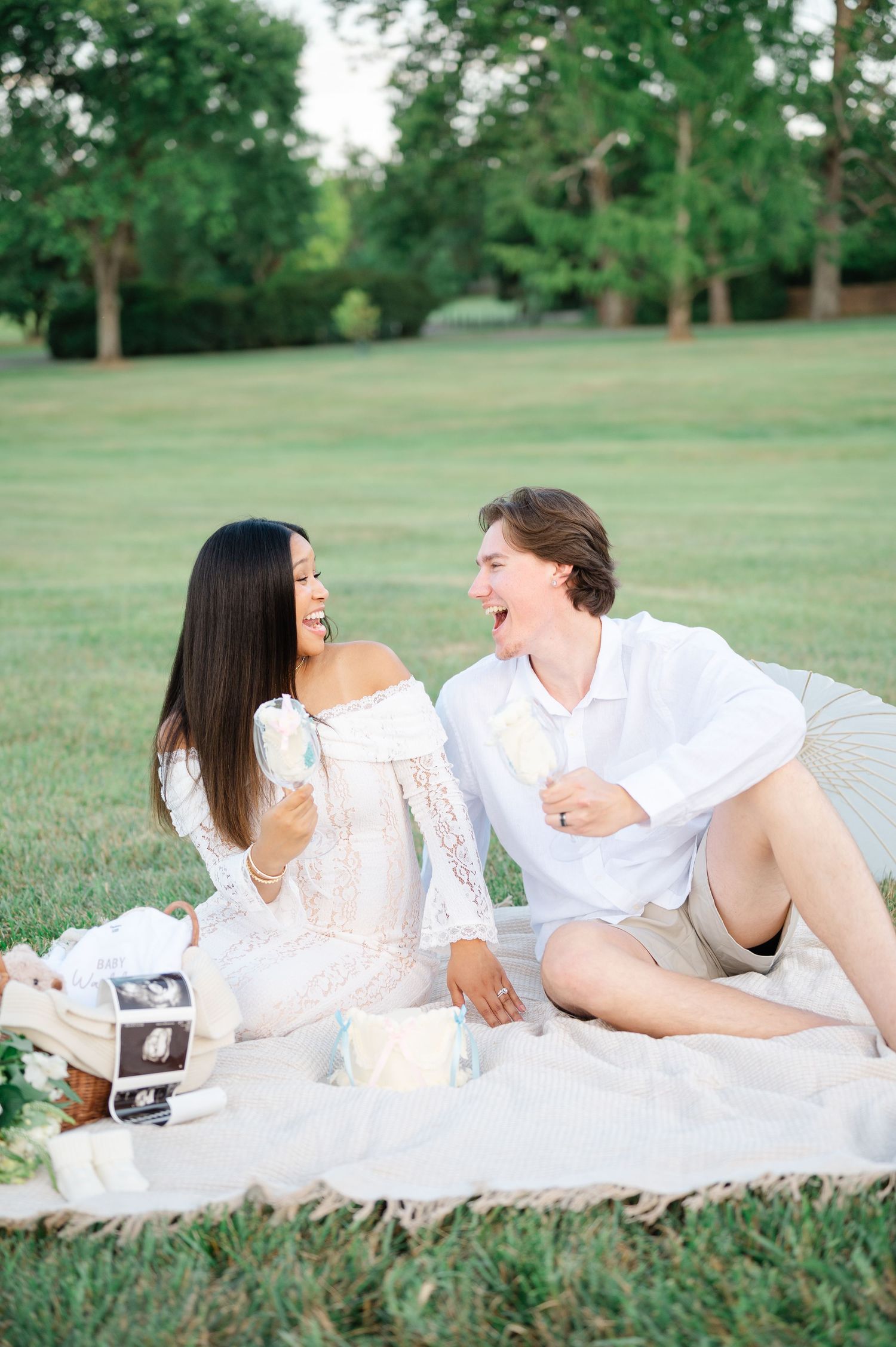 Happy couple in white outfits sharing a picnic on a blanket with ultrasound photos, celebrating pregnancy outdoors.