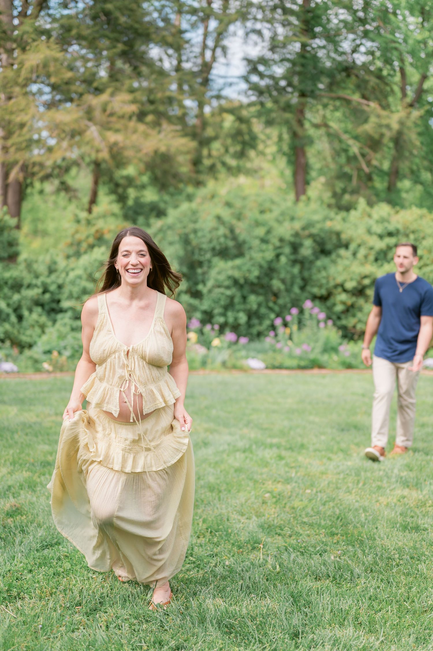 A laughing woman in a yellow ruffled maxi dress walks through a lush green garden as a man follows behind her.