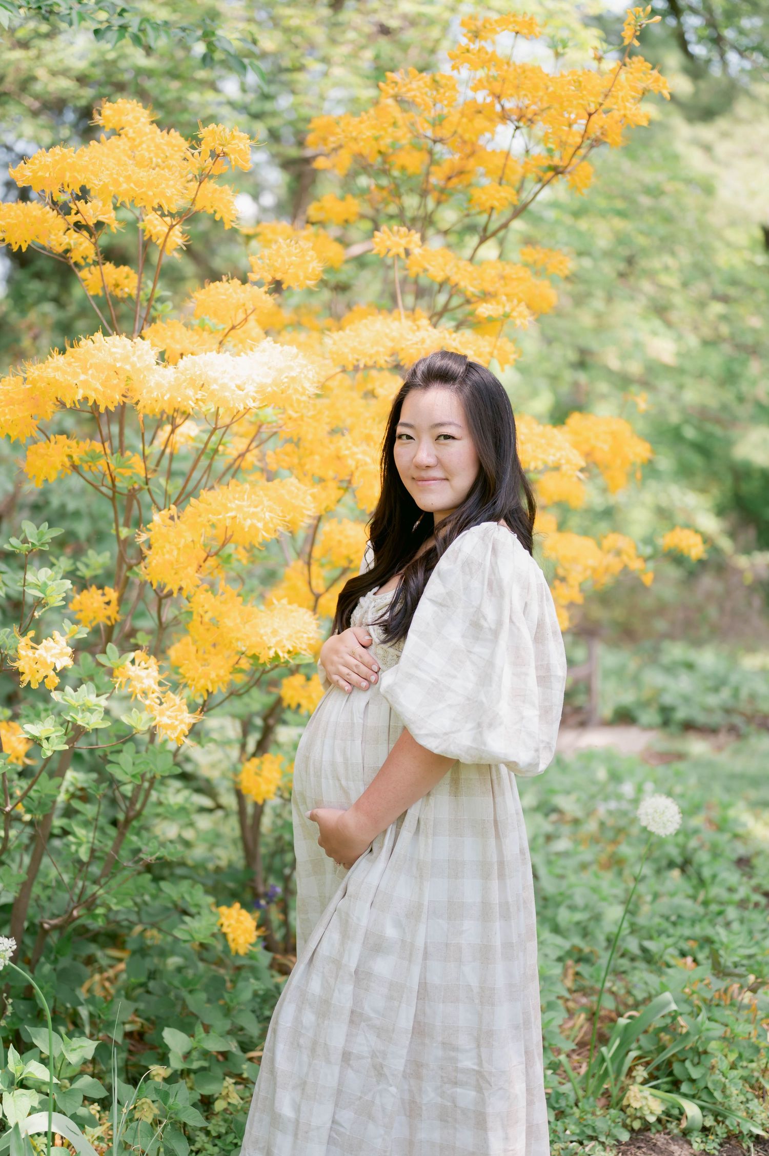 Pregnant woman in white dress holding belly beside yellow flowering shrubs in a lush garden setting.