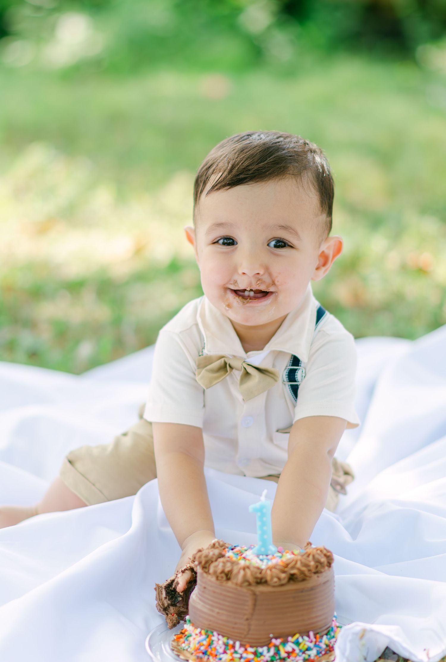 A joyful baby in a white shirt and colorful bloomers plays on a white blanket in a sunny outdoor setting.