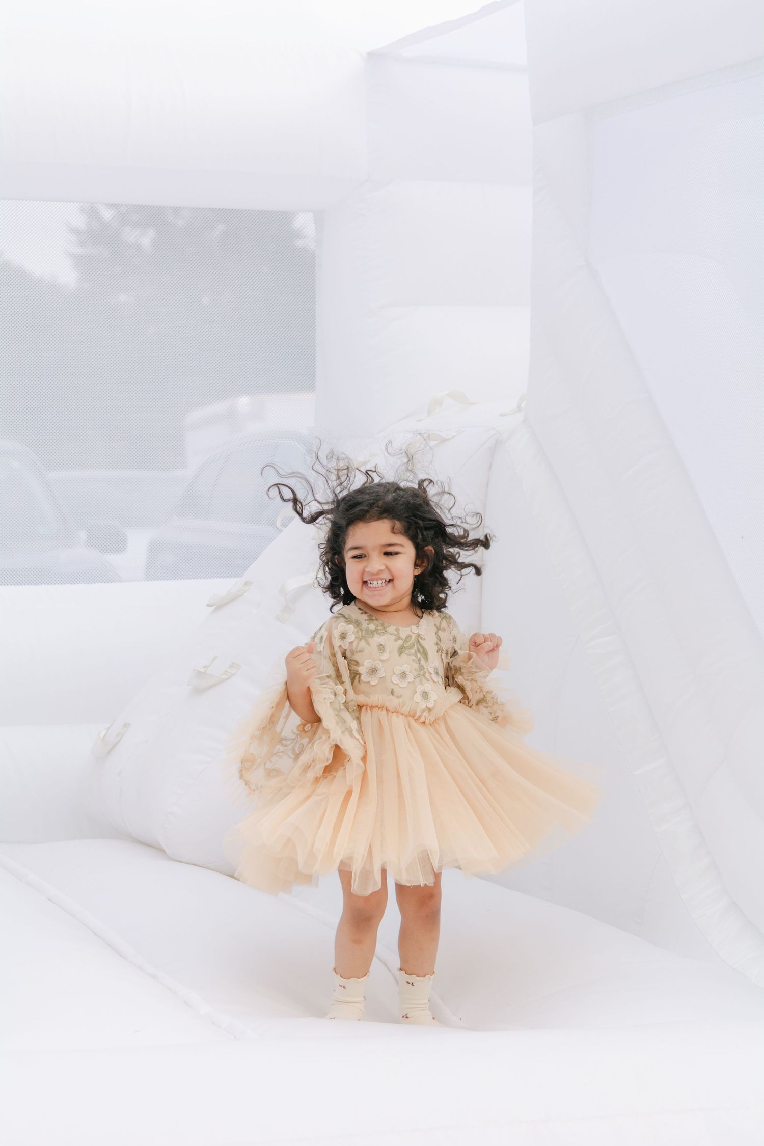 A toddler in a flowing peach dress twirls and plays against a bright white background.
