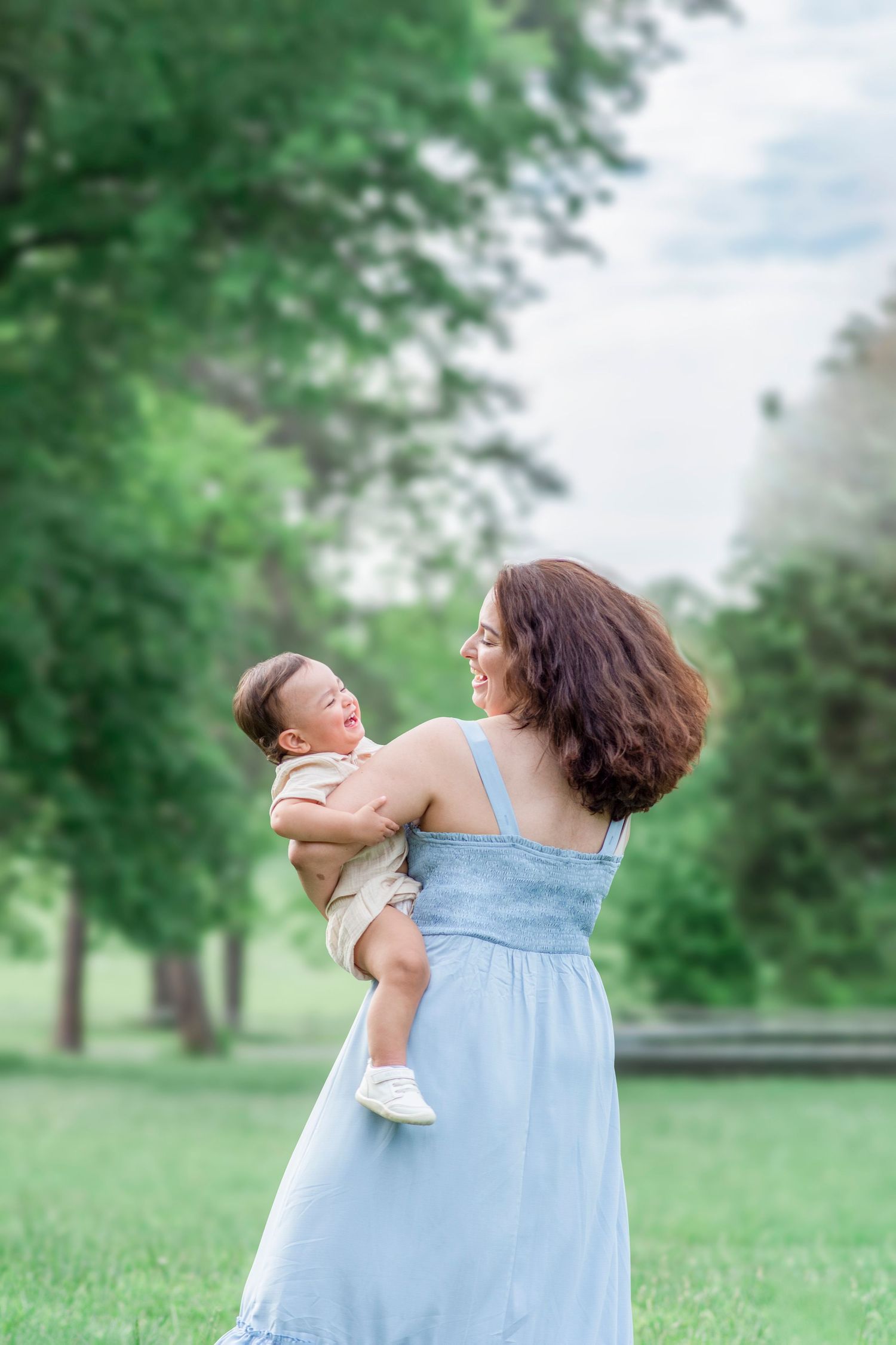 Parent wearing blue dress spins joyfully with baby in sunlit park setting.