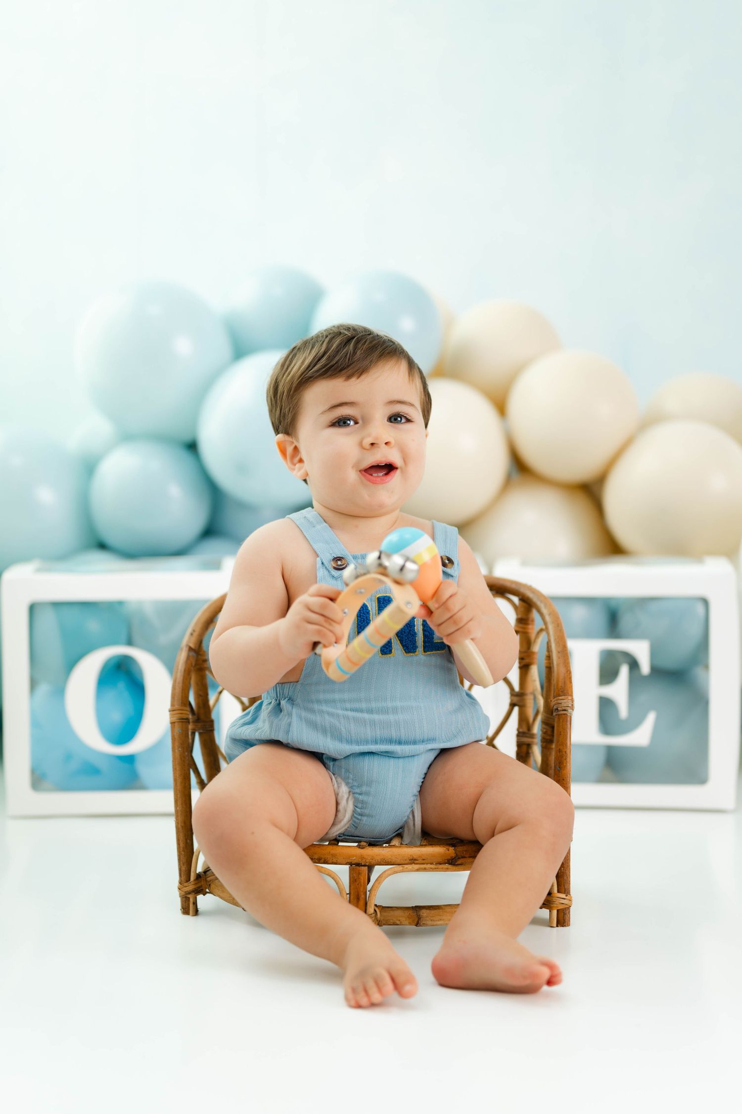 Baby in blue romper sits on wooden chair with balloon decorations in soft pastel colors in background.