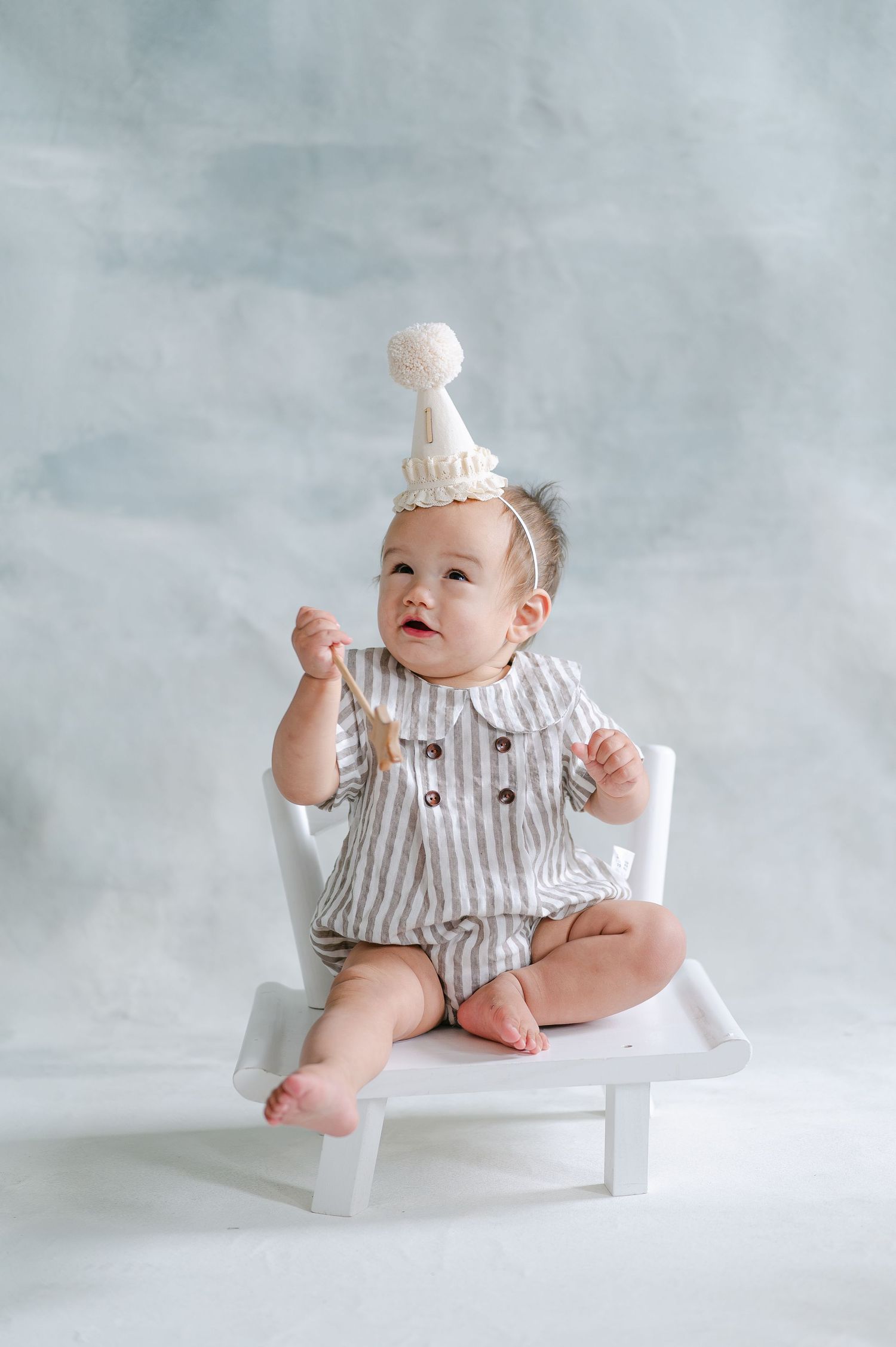 Cute baby in striped romper and white party hat sitting on small white chair against gray background.
