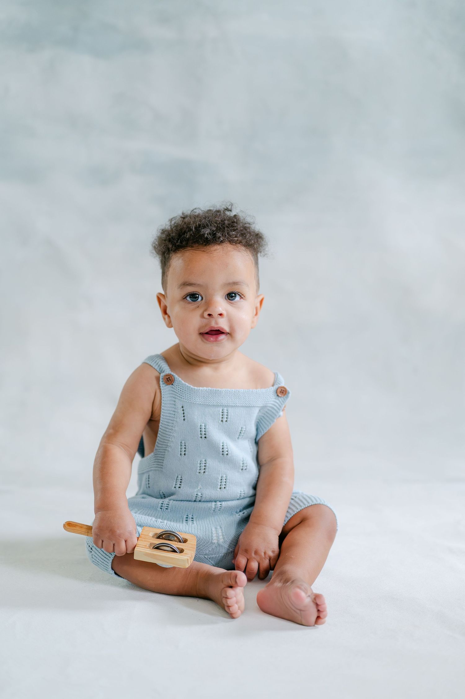 Adorable baby in blue knit romper sitting on white background holding a wooden toy.