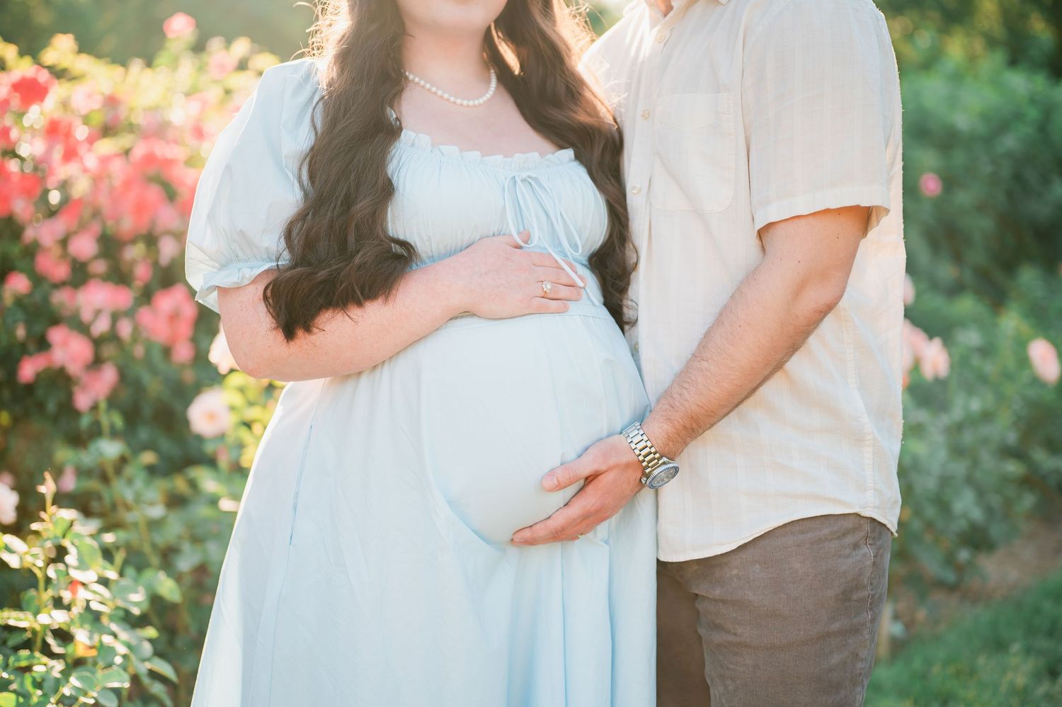 Pregnant couple posing outdoors in a garden with pink roses, woman in light blue dress cradling baby bump.